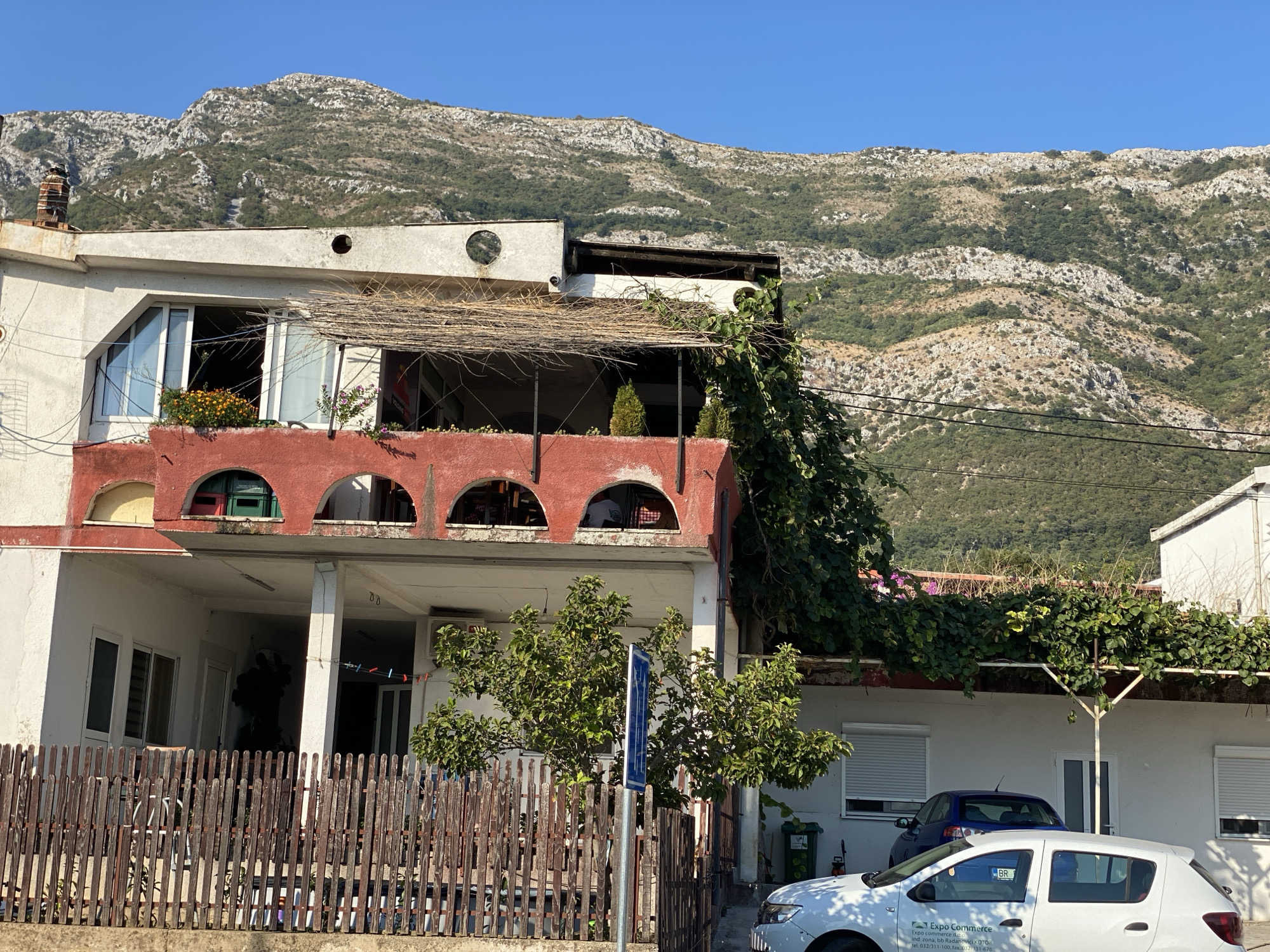 A sunny day in Zgrade, Montenegro, showcases a multi-story white and red building with arched windows and a straw-covered balcony. A lush green mountain range serves as a backdrop. In the foreground, a wooden fence encloses a small garden with a tree and a blue sign. A white car with an "Expo Commerce" logo is parked in front of the building, alongside a blue car. The scene is characterized by clear blue skies, visible power lines, and residential architecture.