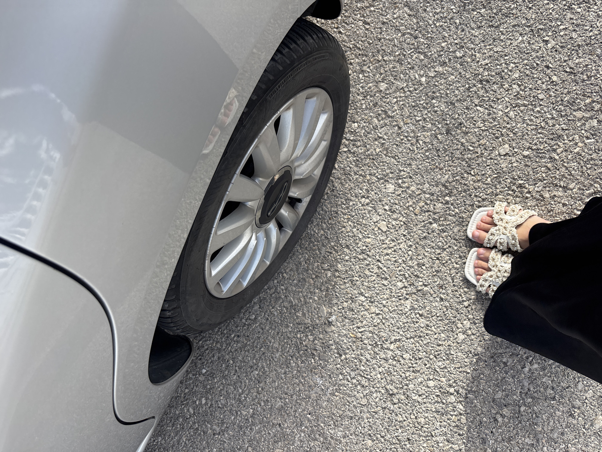 The image shows a close-up, eye-level shot of a person's feet wearing white, ornate sandals standing on asphalt. The person is wearing a black garment that extends down from their legs. To the left of the feet is the rear wheel and lower body panel of a silver car, positioned on the same asphalt surface. The car's wheel features a silver rim with multiple spokes. The scene appears to be outdoors, likely in a street or parking area. The image does not contain any recognizable landmarks or specific features that would pinpoint its location within Orihuela, Spain.