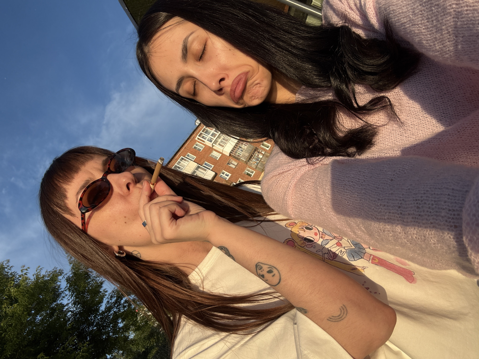 A casual, tilted outdoor photo captures two young women under golden hour light, suggesting late afternoon or early evening.
In the foreground, a woman with brown hair and bangs wears sunglasses on her head. She is holding a hand-rolled cigarette to her lips, inhaling or exhaling. She wears a white t-shirt, and several tattoos are visible on her right arm: a small cartoon alien face and a small rainbow. A silver ring with a dark stone adorns her left pinky finger, and a peace sign tattoo is visible near her left ear.
Behind her, another woman with long dark hair wears a light pink knitted sweater. Her eyes are closed, and she makes a playful, pouting facial expression. Her arm is visible, partially covering a white garment with a colorful cartoon print, depicting a character resembling Sailor Moon.
The background features a clear blue sky with faint clouds, a multi-story residential building with numerous windows and balconies, and lush green trees illuminated by the warm sunlight. The overall scene conveys a relaxed outdoor moment in Votkinsk, Russia. No discernible text is visible in the image.