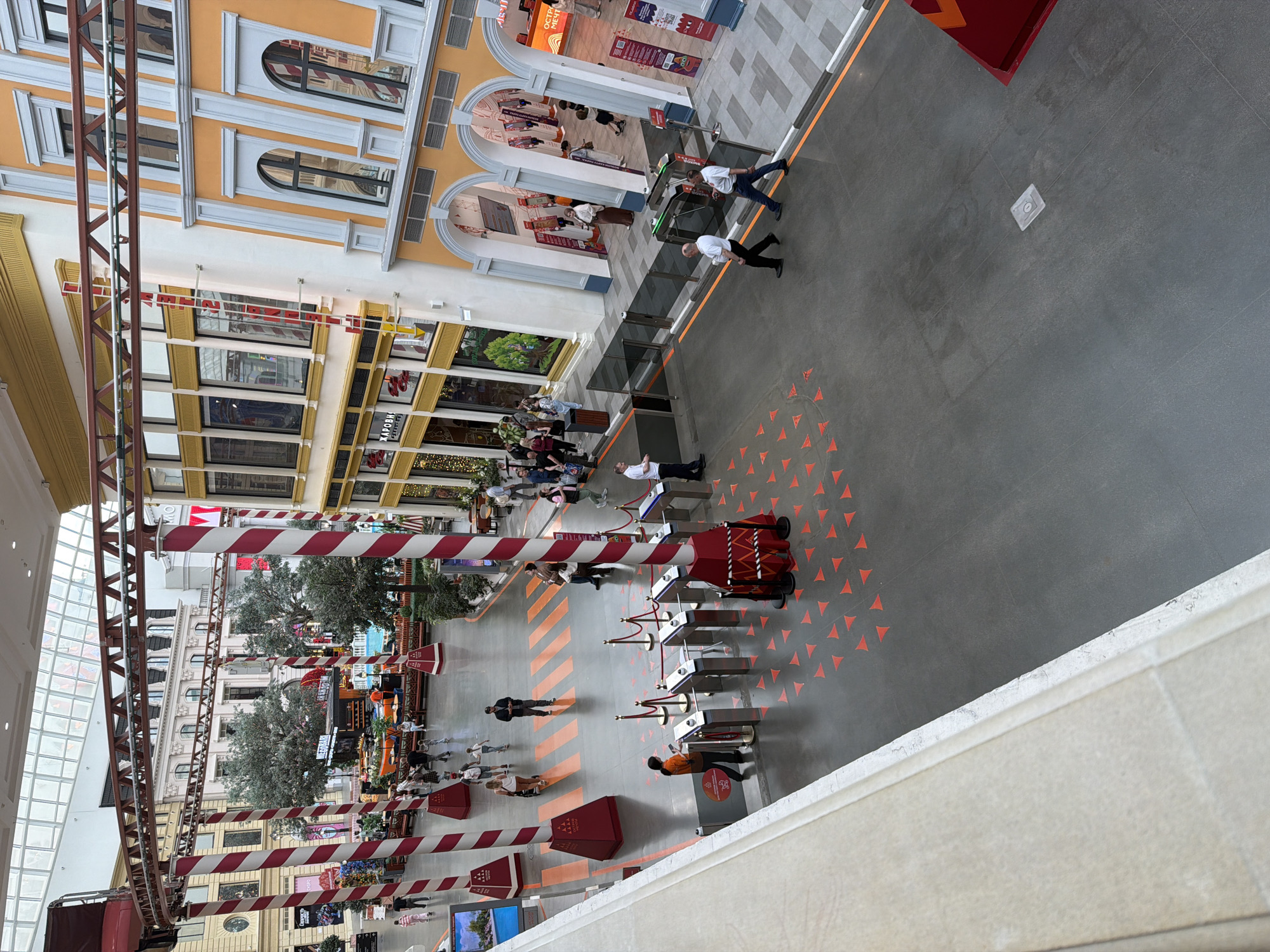 This is a high-angle, full shot of an indoor entertainment venue, likely an amusement park or a themed shopping mall. The scene is vibrant and colorful, with a focus on a walkway that transitions into a play area. Several candy-cane striped pillars, typical of a festive or whimsical theme, punctuate the space.
In the foreground, a section of red-and-white striped pillars and an overhead track suggest the presence of a ride, possibly a roller coaster. A pathway in the middle ground is marked with orange triangular patterns on a gray floor, guiding pedestrian traffic. Several groups of people are visible, some walking, others standing.
To the left, a building facade with large windows and architectural detailing resembling European city buildings can be seen. In the center, a more relaxed area with trees, seating, and a small pool suggests a leisure zone. Further to the right, another building facade features arched entrances and windows, with more shops or attractions visible behind them.
The overall impression is one of a bustling, family-friendly entertainment destination, with distinct themed zones and clear pathways for visitors.
