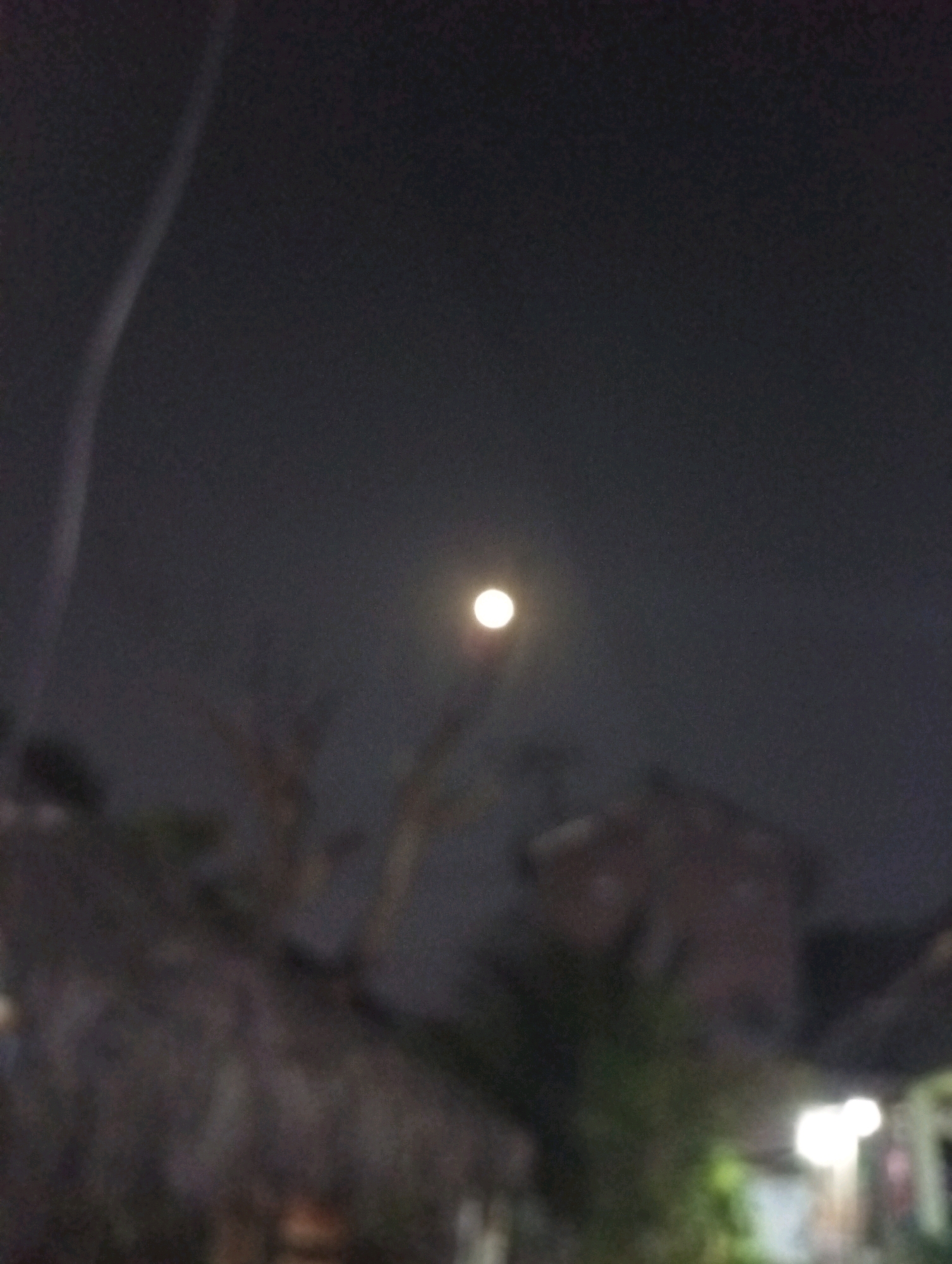 The image shows a nighttime scene in Kediri City, Indonesia. A bright, circular object, presumably the moon, is visible in the dark sky, slightly off-center to the right. A faint, curved streak of light, possibly from a streetlamp or a thin cloud illuminated by artificial light, extends from the upper left quadrant of the image downwards. The lower portion of the image is blurred and depicts indistinct shapes of buildings and foliage, suggesting the photograph was taken from ground level looking upwards. The overall atmosphere is dark, with limited visibility.