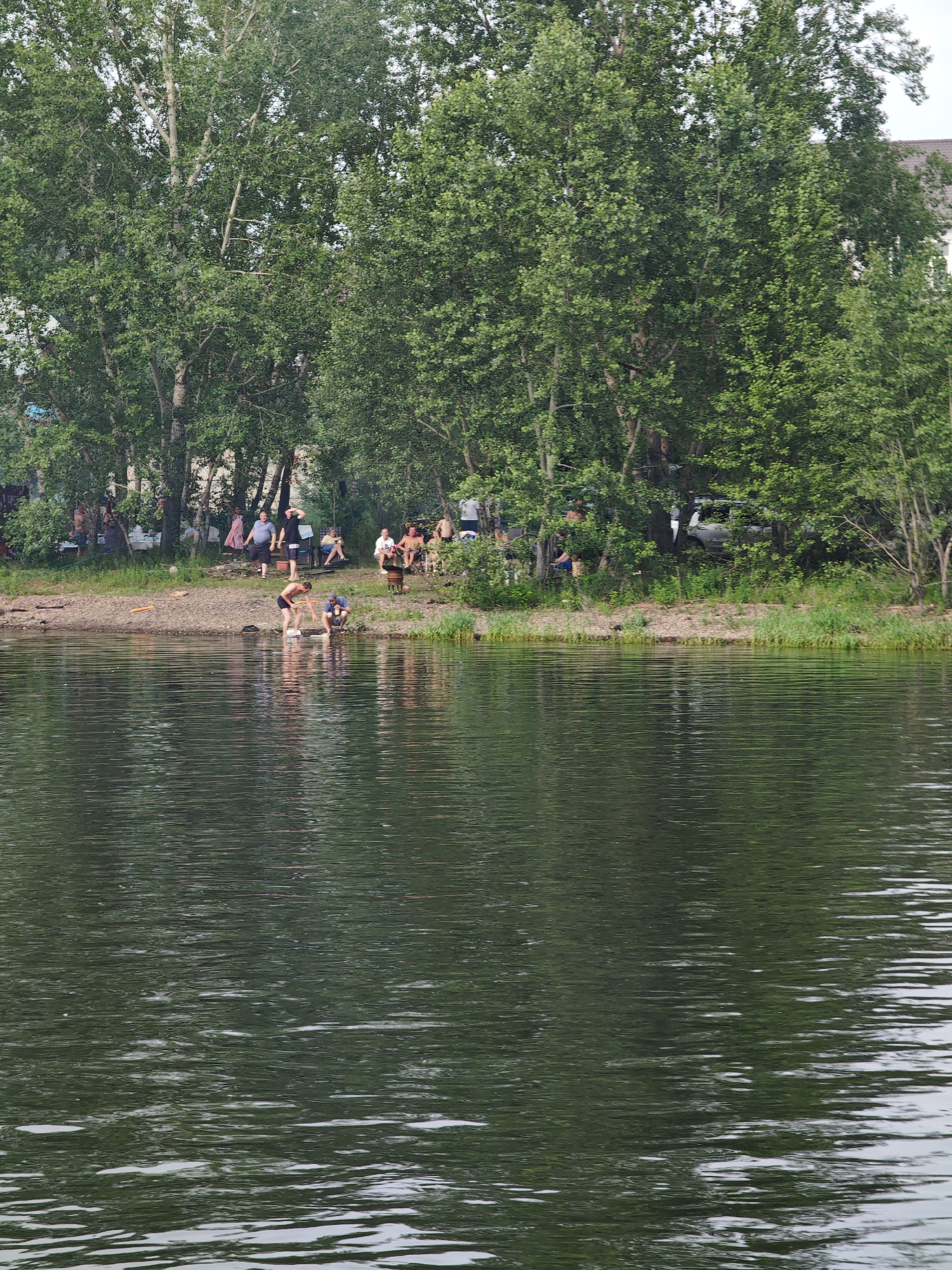 The image depicts a riverside scene in Ермолаевский Затон, Russia. On the grassy bank, several people are gathered under the shade of tall green trees. Some are sitting on chairs or the ground, while others are standing or walking. A few individuals are near the water's edge. In the foreground, the dark, rippling surface of the water fills the frame. The overall atmosphere suggests a casual gathering or outing by the river.