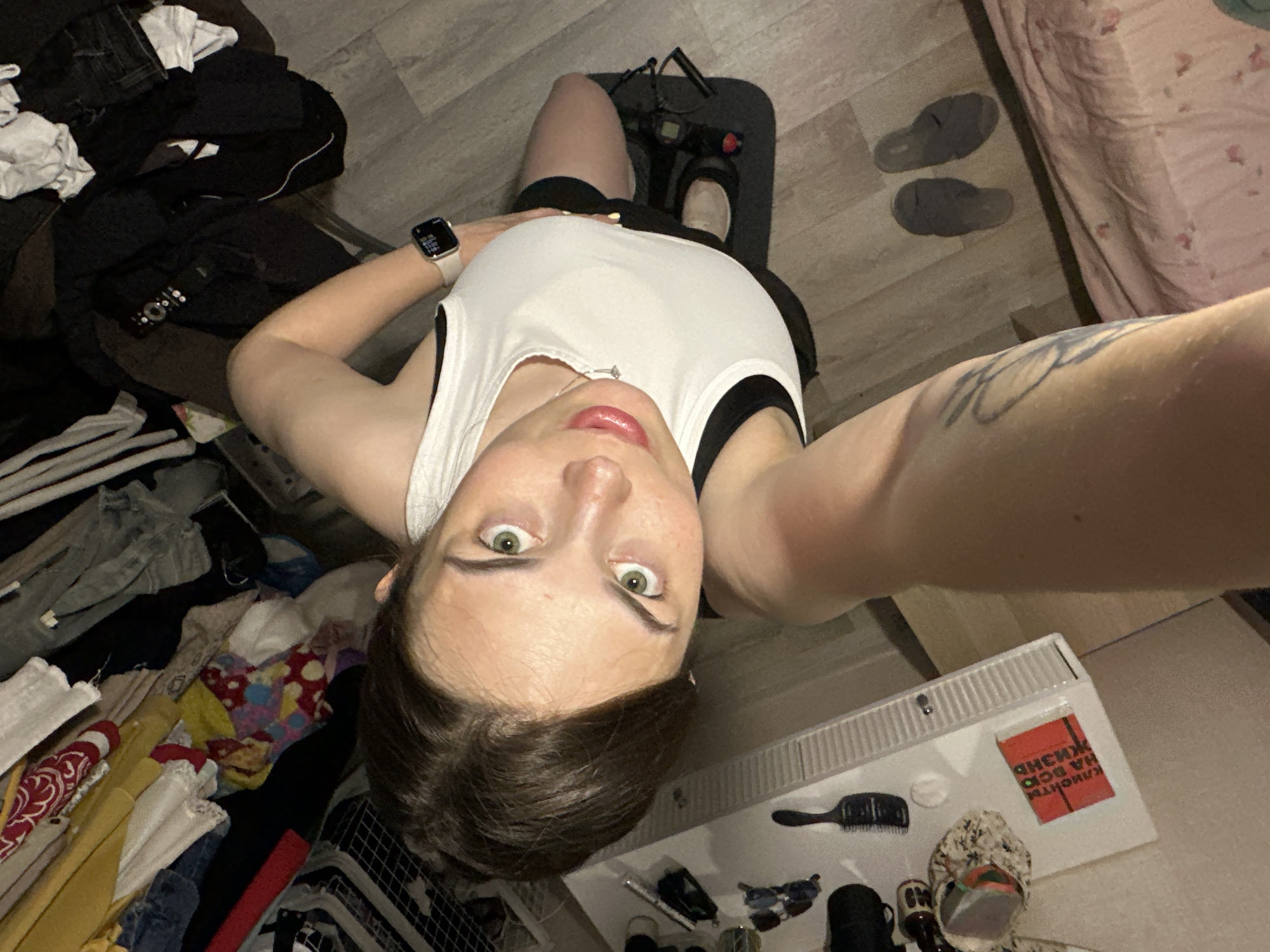 In a domestic setting, a woman with brown hair and green eyes is taking a selfie. She is wearing a white tank top with black trim and black athletic shorts. She has a diamond pendant necklace and an Apple Watch on her left wrist. The background appears to be a room with clothing strewn about, suggesting a closet or laundry area. A white shelf unit with various items, including a red book with Russian text, is visible. The floor is wood laminate. A pair of gray slippers is on the floor to the right of the woman. The image is taken from a high angle, looking down at the woman. There is no explicit indication of the city of Perm, Russia, in the image itself.