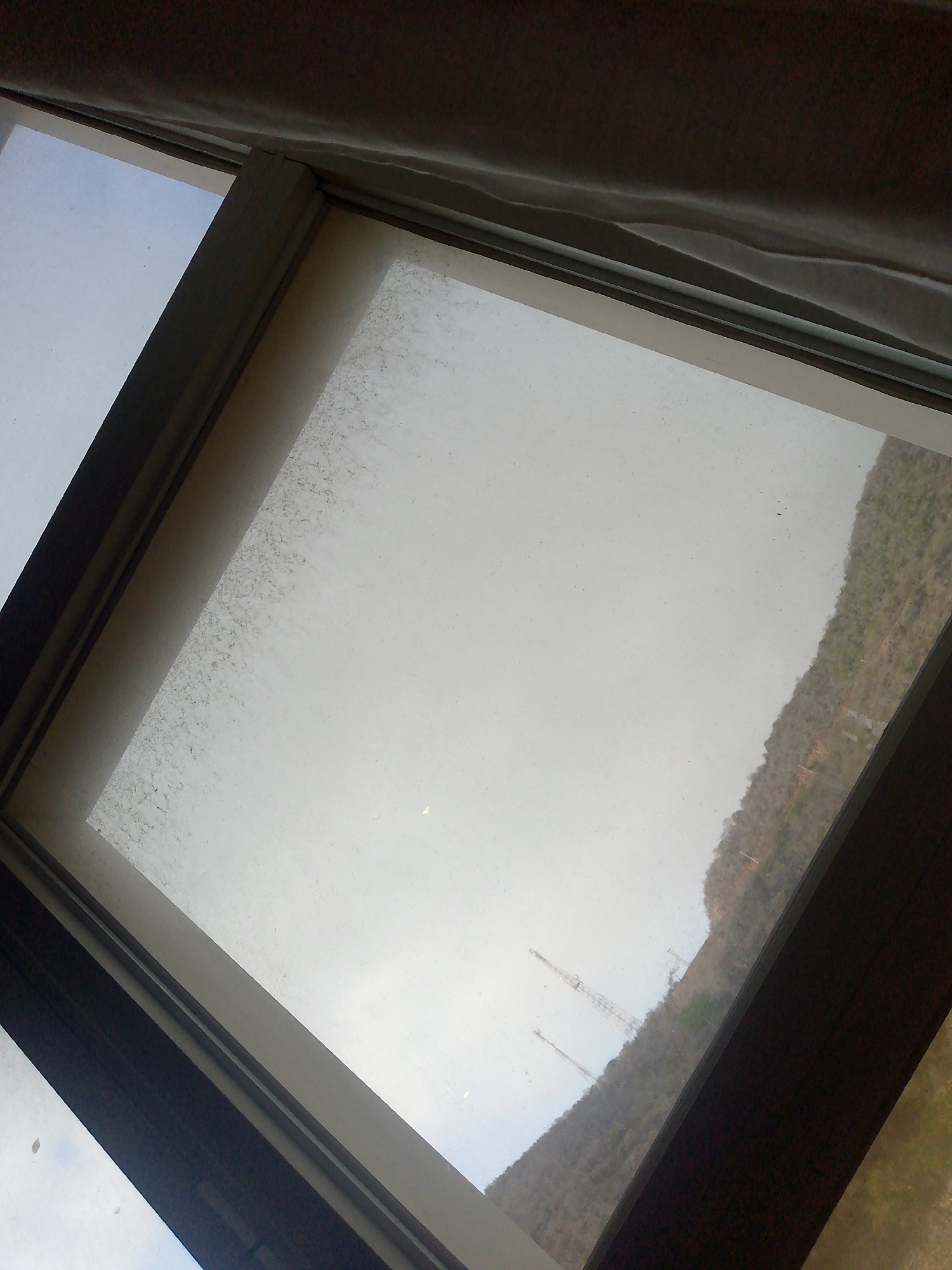 Here's a description of the image:
The image shows a view through a dirty window. Part of a hillside is visible, with a few communication towers on the slope. The sky appears overcast. The window frame is visible, along with part of a curtain or drape at the top. The photo is taken at an angle, looking up and to the side. Given the prompt instructions mentioning Pedro González, Venezuela, it can be inferred that the hillside scene is located there.