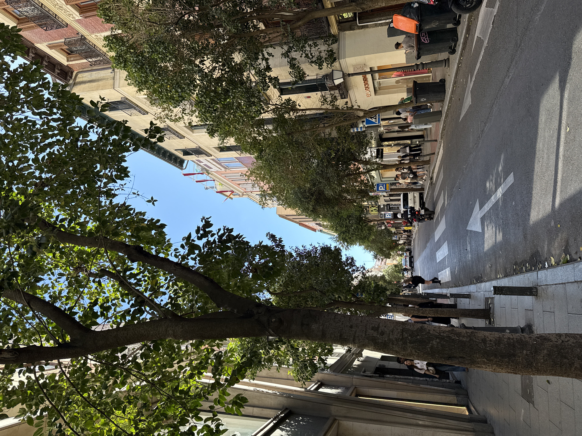 The image shows a street scene in Madrid, Spain. The view is oriented along the street, with buildings on either side. Trees line the street, obscuring some of the buildings. People are walking on the sidewalks and crossing the street. There is street signage and trash cans visible. The sky is clear and blue.