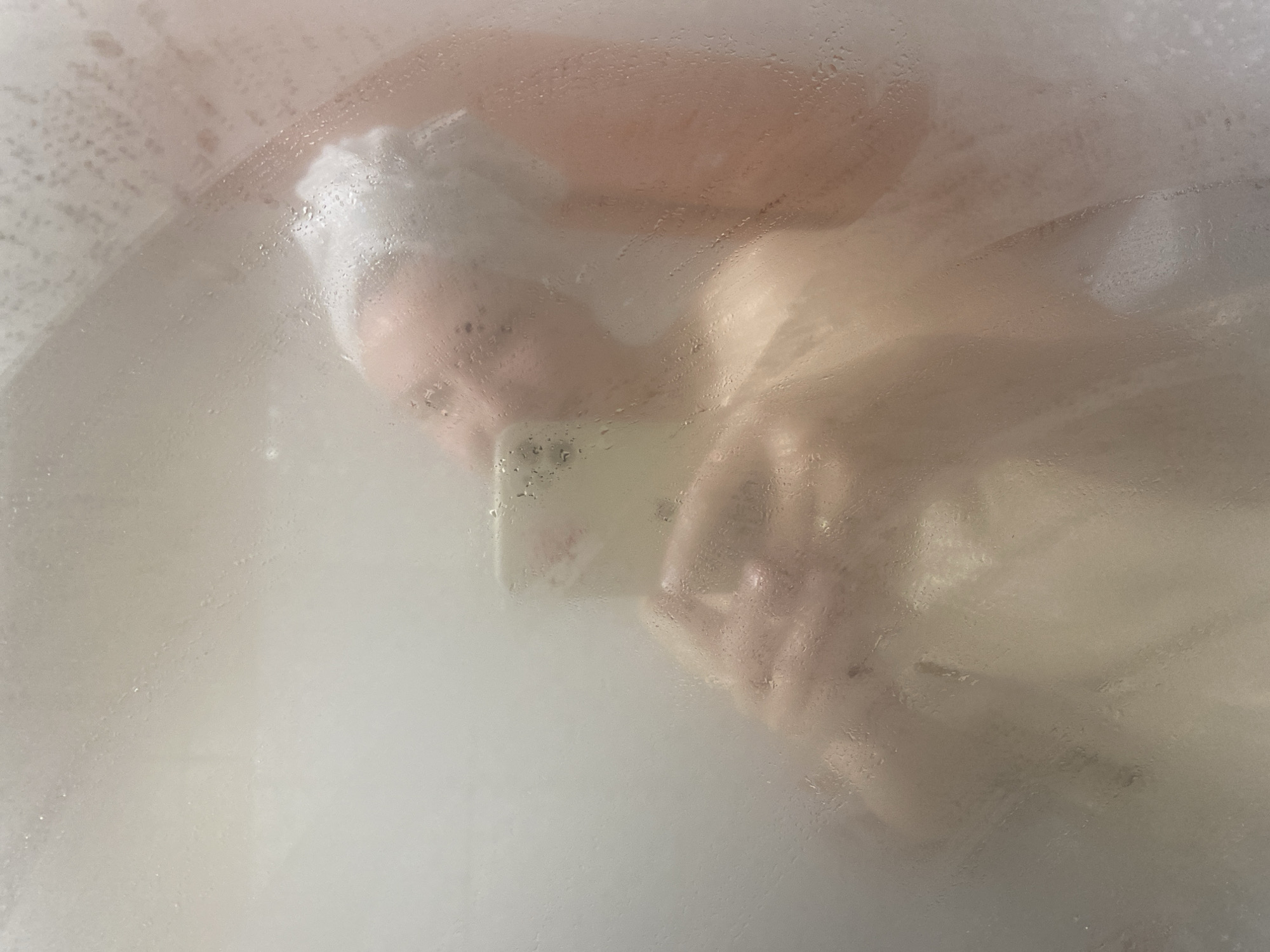 A person is seen behind a foggy or steamy surface, possibly a shower door. The person appears to be holding a cell phone, possibly taking a selfie. They have a white covering on their head, likely a shower cap or towel. Details are obscured by the condensation. The location in Chaikovsky, Russia, cannot be determined from the image alone.