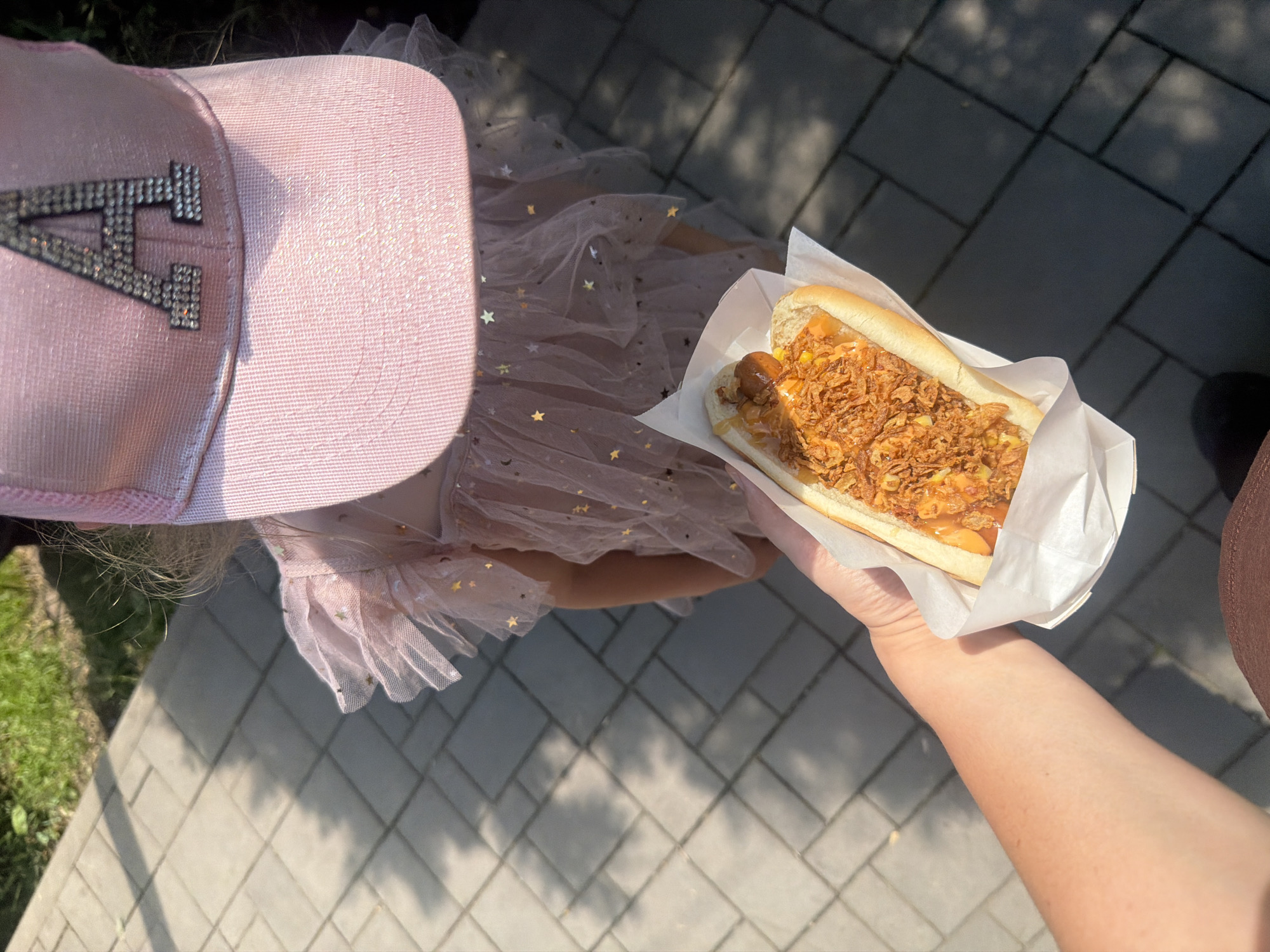A person's arm and hand, holding a hot dog in a white paper wrapper, extend into the foreground. The hot dog is in a bun and topped with cheese, sauce, and crispy fried onions. In the background, a young child is visible from the shoulders up, wearing a pink baseball cap with the letter "A" adorned with rhinestones. The child is also wearing a light pink, sparkly dress. The scene appears to be outdoors, with a paved walkway made of grey rectangular blocks and a patch of green grass in the upper left corner. Sunlight casts shadows on the ground. The location is described as Unknown, Unknown, in the city of Unknown.