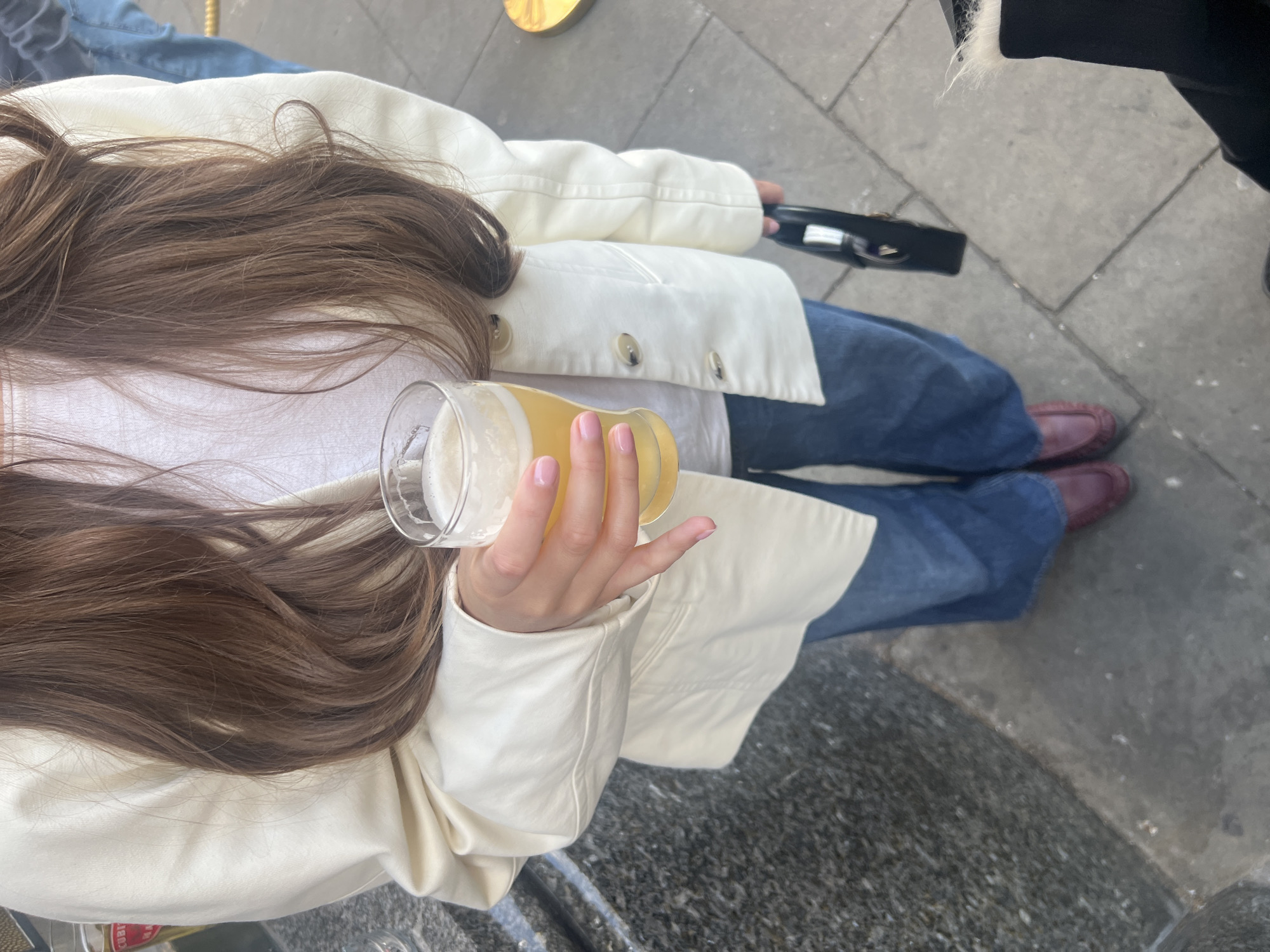 The photograph shows a person from an overhead perspective, holding a glass of beer. The person has long, brown hair and wears a white jacket with visible buttons, blue jeans, and maroon-colored loafers. A black purse is also visible in the upper right quadrant. The ground is a grey stone pavement, suggesting an urban setting. Based on the attire and the style of the street, it is potentially located in London, United Kingdom.