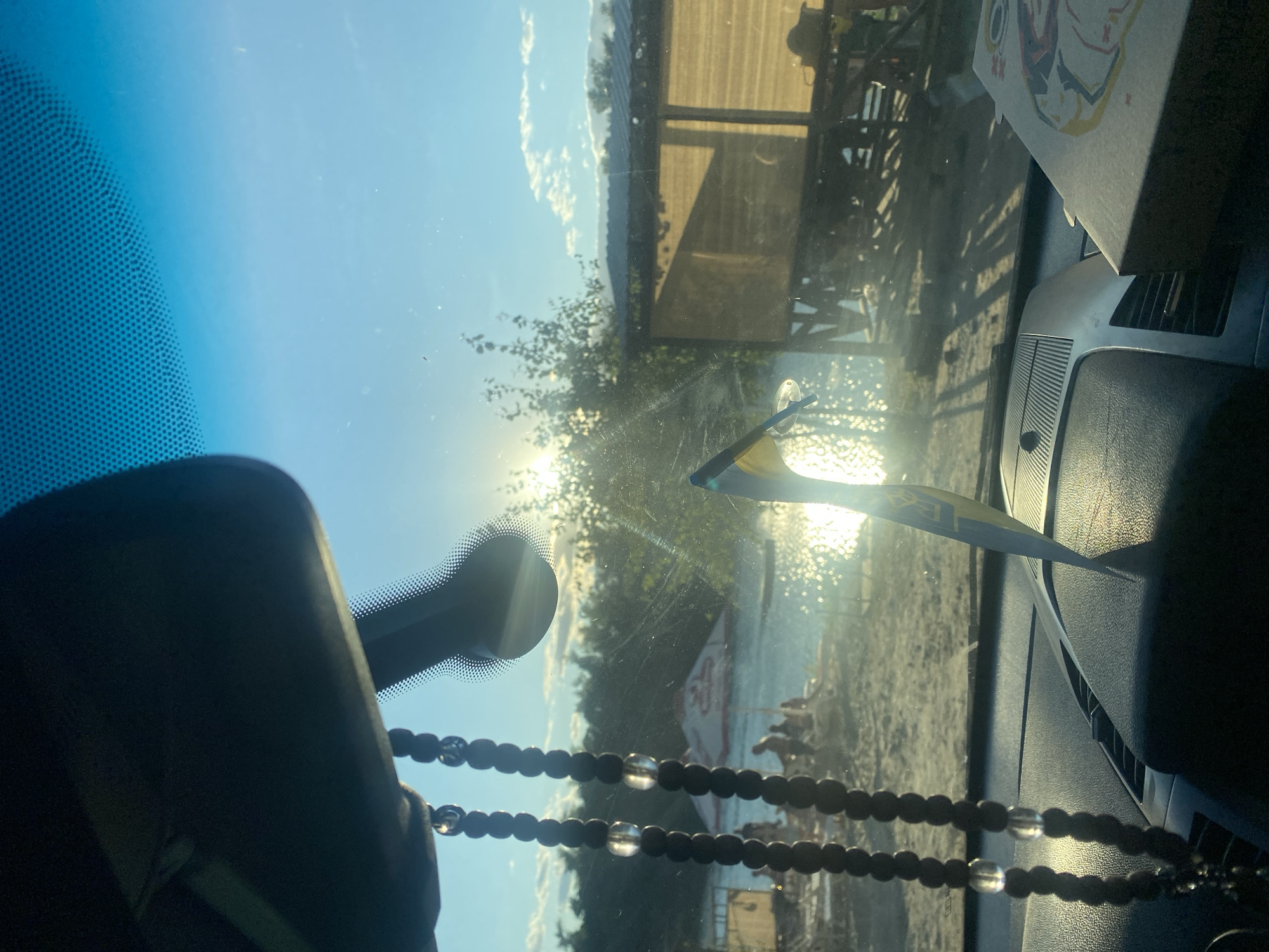 The image is a first-person perspective from inside a car, looking out through the windshield. The sun is shining brightly, causing lens flare and obscuring some details of the outdoor scene. In the foreground, a string of dark prayer beads with clear interspersed beads hangs from the rearview mirror. On the dashboard, a small object, possibly a box with an illustration, is visible on the right.
The view through the windshield shows a beach or lakeside setting. The water is reflecting the sunlight, creating a shimmering effect. On the shore, there are several people, some appearing to be sitting or lying down on lounge chairs or towels. There are also structures on the beach, including what looks like a small wooden building or kiosk with a yellow roof and a red and white umbrella behind the people. Trees and foliage line the background. The sky is blue with some scattered white clouds.
The presence of the Ukrainian flag is implied by a yellow and blue item visible in the center of the windshield, suggesting the location is in Ukraine, specifically Dnipro, Ukraine, as per the prompt. The overall atmosphere suggests a sunny day at a recreational area.