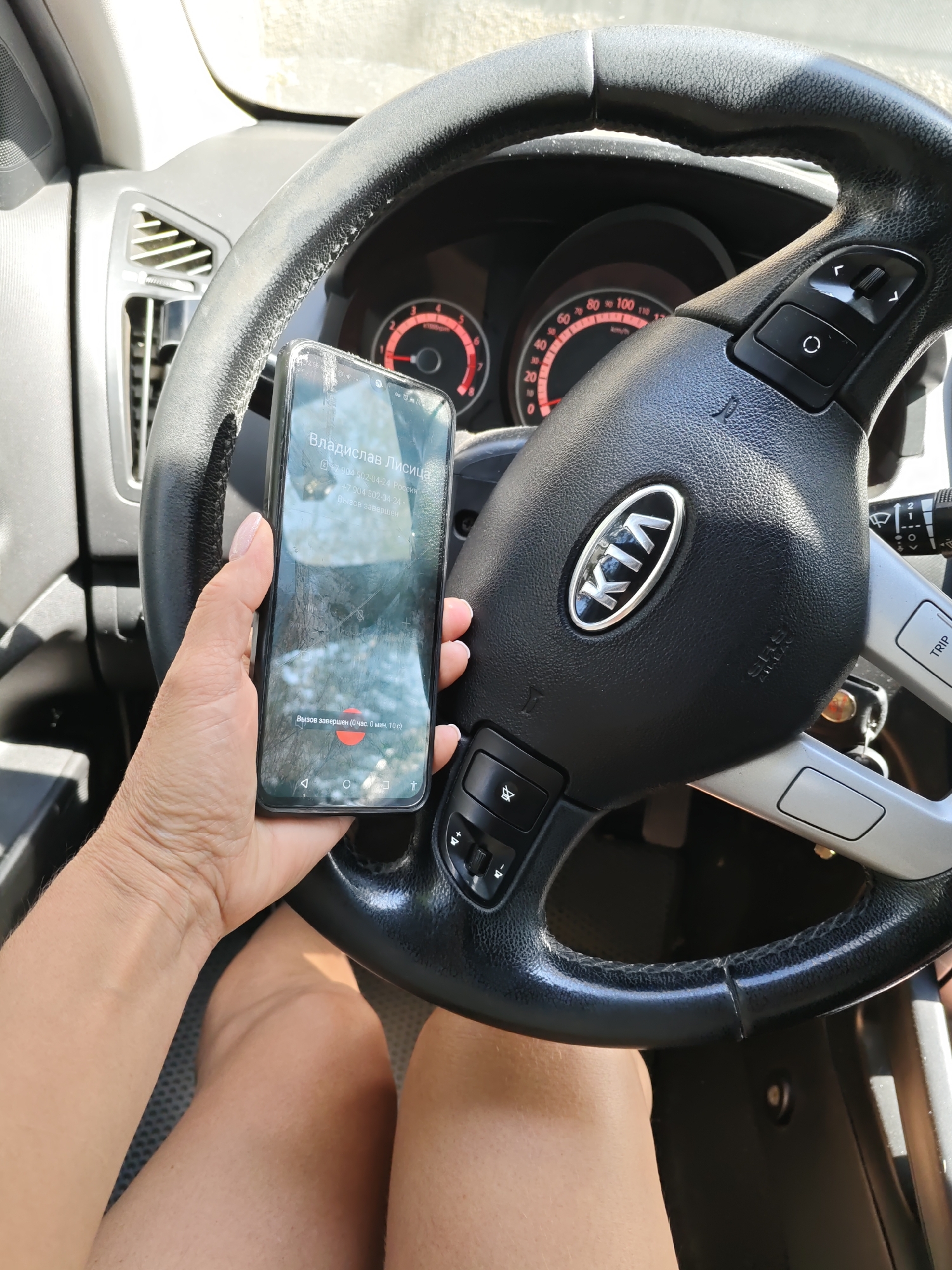 A person's hand, with light skin and short fingernails painted with light pink polish, holds a black smartphone with a cracked screen. The phone displays a call screen showing "Vladislav Lysitsa" and the number "+7 904 502-04-24 Russia". Below that, it indicates "+7 904 502-04-24" and "Call ended". The bottom of the screen shows "Call ended (0 hours, 0 min, 10 sec)" with a red circular icon. The phone is positioned in front of a black Kia steering wheel. The steering wheel has silver accents and buttons, including a Kia logo in the center. The dashboard with red-faced gauges is visible behind the steering wheel. The scene appears to be inside a car, likely in the city of Rostov-on-Don, Russia, given the phone's display of the Russian phone number. Sunlight illuminates the interior of the car.