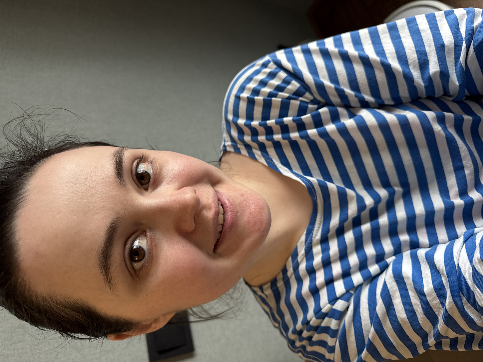 A woman with brown hair and eyes is angled toward the camera, wearing a blue and white striped shirt. The background consists of a gray surface, with a black object visible near the bottom left. The city and location are unknown and cannot be inferred from the given information.