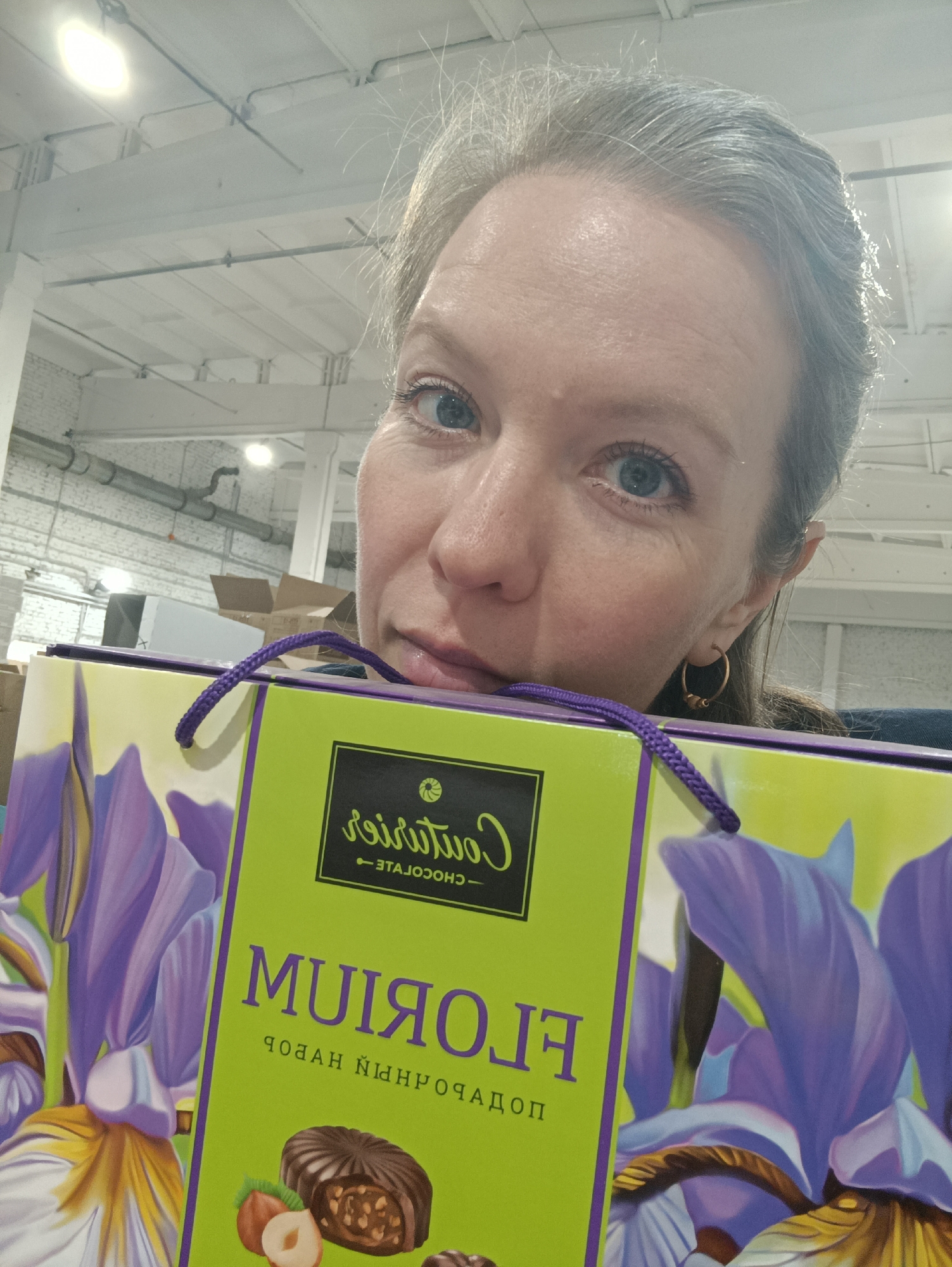 A person with blue eyes and graying hair holds up a box of "Courtois" chocolates in front of a room in Chaikovsky, Russia. The box is decorated with a floral pattern of purple irises on a green background. The label reads "FLORIUM" in Russian, with an image of a hazelnut and chocolate candy at the bottom. The background shows a warehouse-like room with white walls and exposed pipes.