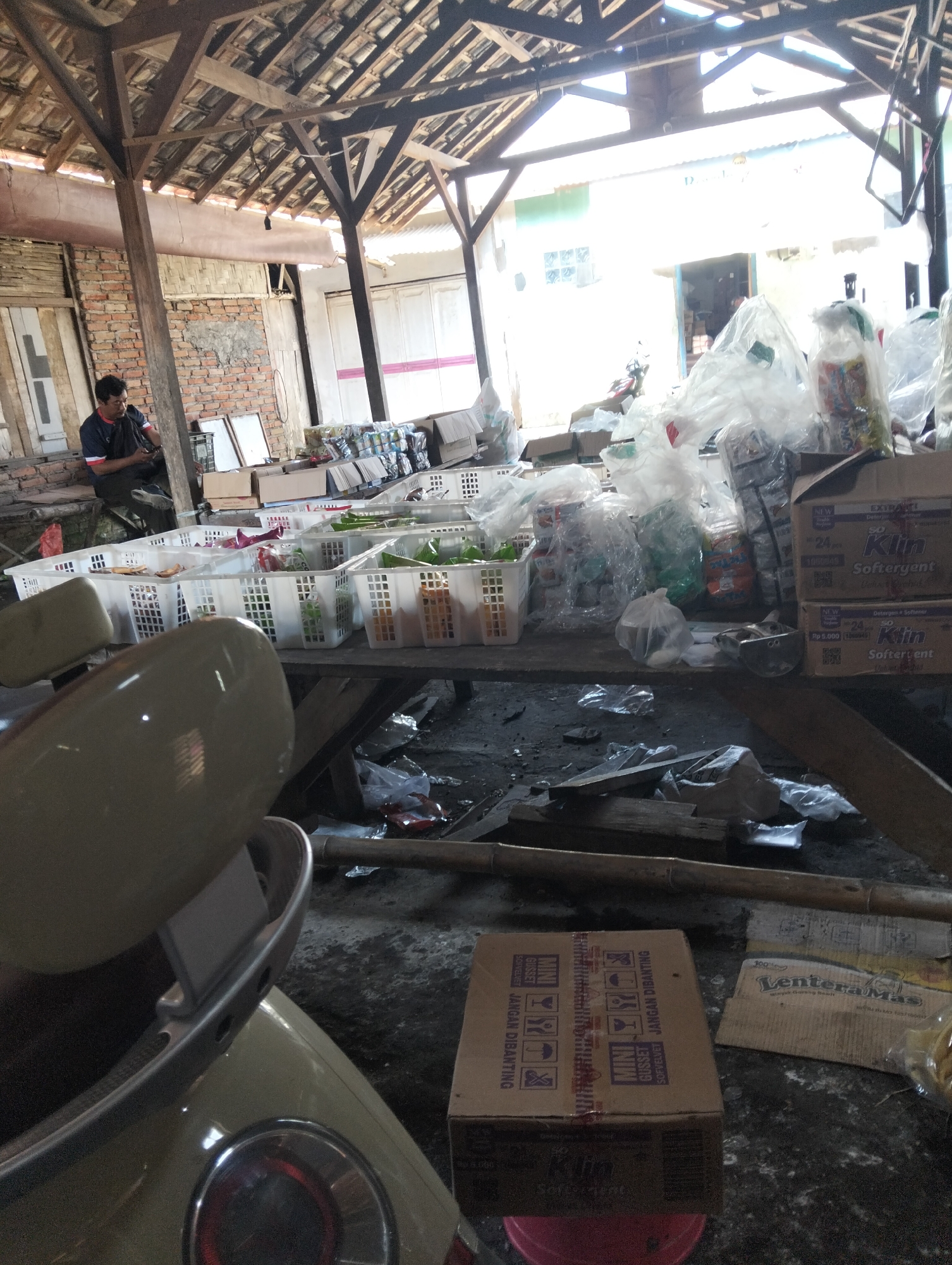 The image depicts an interior scene within what appears to be a warehouse or open-air market stall in Muncar, Indonesia. The structure has a rustic wooden beam ceiling with a corrugated roof, allowing natural light to filter in. In the foreground, the back of a light-colored scooter or motorcycle is visible.
A man wearing a dark blue shirt with red and white stripes on the sleeves and dark pants is seated on a low surface to the left of the frame, looking down at his mobile phone. He is positioned near a brick wall with bamboo detailing.
The central area of the scene is dominated by a long table laden with numerous white plastic baskets filled with various packaged goods. These goods include detergent boxes (brand "So Klin" is visible), plastic bags of what appear to be snacks or food items, and other miscellaneous packaged products. Some of these baskets are partially covered with clear plastic sheeting.
Behind the table, stacks of cardboard boxes and more packaged goods are visible, some of which are also covered in plastic. The floor is uneven and appears to be made of concrete or dirt, with scattered debris including plastic bags, pieces of wood, and discarded packaging. The overall impression is one of a busy, functional storage or retail space.