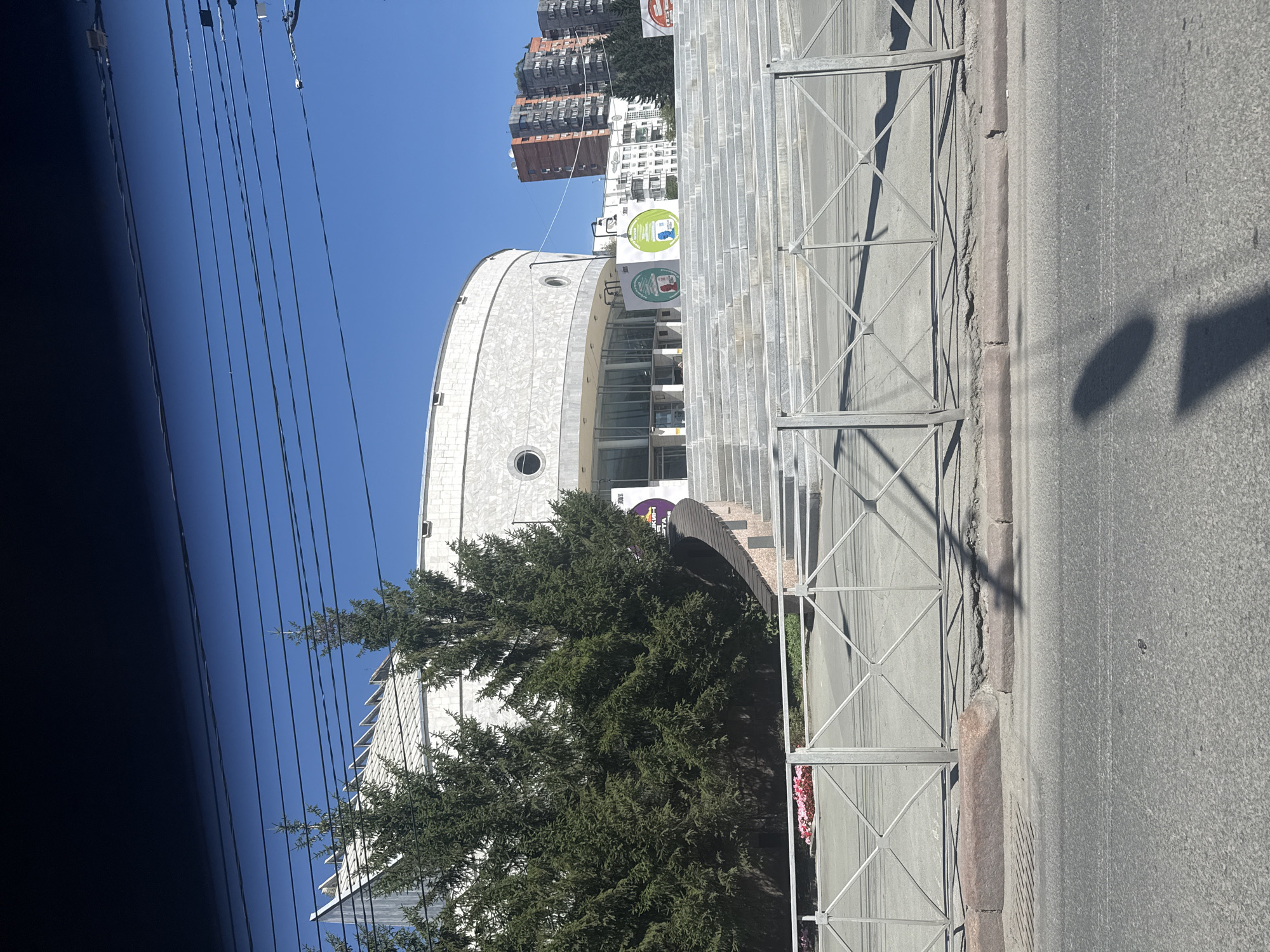 The image captures a wide, sunlit view of the Novosibirsk State Academic Opera and Ballet Theatre, commonly known as the Novosibirsk Opera and Ballet Theatre. The theatre, a prominent landmark, is characterized by its large, semi-circular facade constructed of light-colored stone or concrete. Its distinctive roofline features a series of sharp, angular peaks. In front of the theatre, a grand, wide staircase made of stone ascends towards the entrance, flanked by dark green conifer trees on the left. Several large, circular posters are visible on display near the base of the stairs. A metallic fence runs horizontally across the foreground, separating the theatre's grounds from a paved street. Beyond the theatre, multi-story residential buildings and power lines are visible against a clear blue sky. The scene is devoid of any visible individuals.