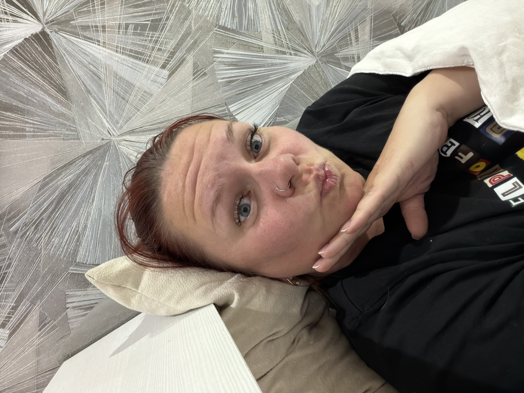 The image captures a close-up, angled view of a person lying down indoors, likely in Chaikovsky, Russia. The subject is a female with reddish-brown hair, blue eyes, and a silver nose ring on her left nostril. She wears a black short-sleeved t-shirt featuring fragmented, multi-colored text and shapes, including partial letters like "C", "U", "T", "E", "P", "O", "F", and "R". A white blanket or duvet is draped over her upper body and right arm.
Her head rests on a light beige or off-white pillow, and a light-colored, possibly wooden or laminated, surface is visible next to it in the foreground. She is looking directly at the camera with a playful expression, her lips pursed in a "kissy face." Her left hand is raised, with fingers gently touching her chin, revealing manicured nails with white tips.
The background is a prominent feature: a silver and grey patterned wallpaper with an abstract, geometric design composed of radiating lines and angular shapes, creating a textured and dynamic visual. The overall scene suggests a casual, relaxed moment, possibly a selfie taken in a personal indoor setting.