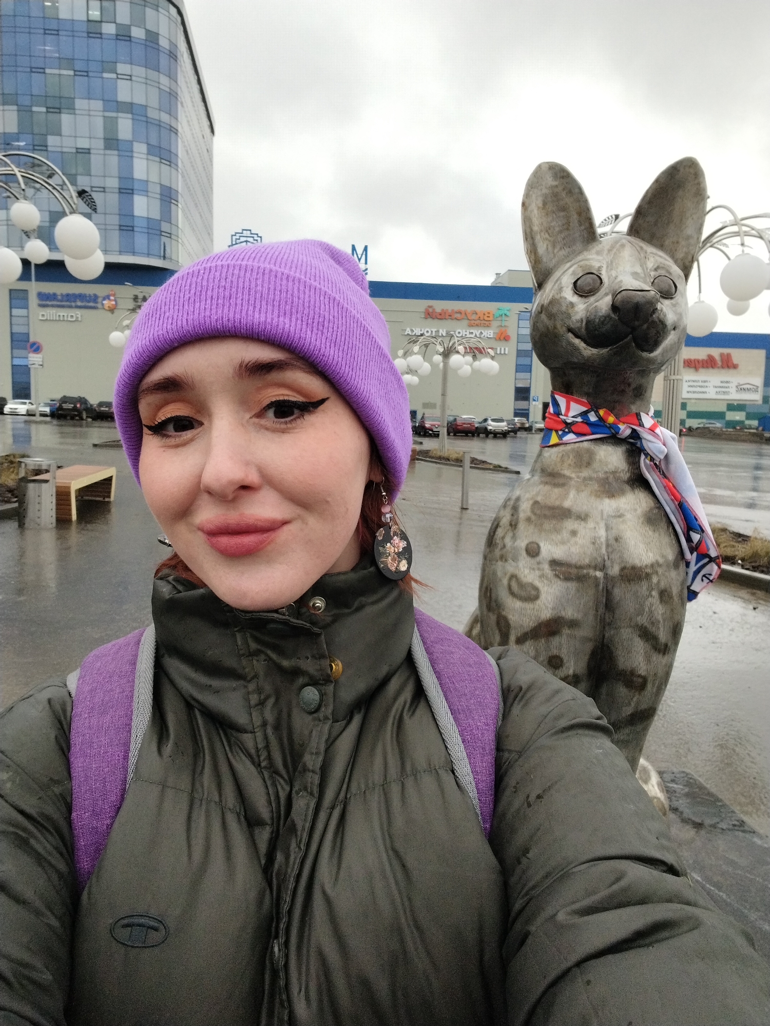 A woman is taking a selfie next to a metallic dog statue wearing a colorful scarf. The woman has red hair, fair skin, a purple beanie, and a dark green puffer jacket with a purple backpack strap visible. She is wearing earrings and makeup, including winged eyeliner and lipstick.
Behind the woman and the statue is an urban landscape with a large, modern building. The building has blue tinted glass on its upper floors and advertising signage on its lower floors. Further in the background, cars are visible on the wet pavement.
The setting appears to be a rainy day based on the wet ground and the overcast sky. The scene is located in Izhevsk, Russia.