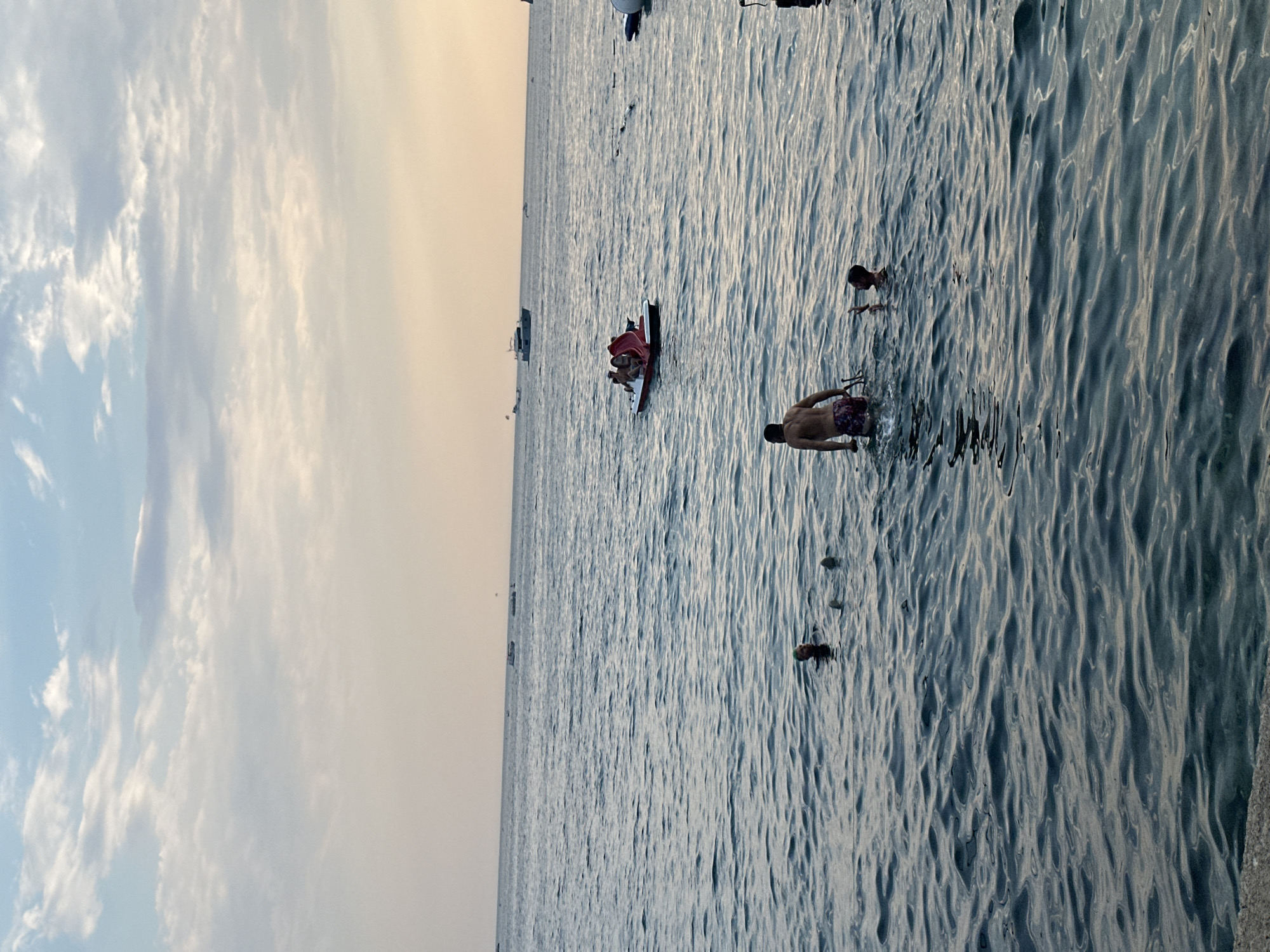 An outdoor, eye-level, long shot captures a serene scene at sea, likely near Budva, Montenegro, under a hazy, pale blue sky with scattered clouds. The ocean, a deep, rippling blue, dominates the frame, reflecting the muted light of what appears to be late afternoon or early evening.
In the foreground, several individuals are engaged in water activities. Two people, their heads barely above the water, are situated to the left. Further right, a man with his back to the camera, wearing patterned swim shorts, stands waist-deep in the water, holding a pair of metal poles. To his right, a child is also in the water, with their right arm raised.
Further out at sea, a small red and white pedal boat carries four people, two adults and two children. Beyond this, several boats are visible on the horizon, including a larger white vessel. The overall impression is one of leisurely enjoyment of the water and the coastal environment.