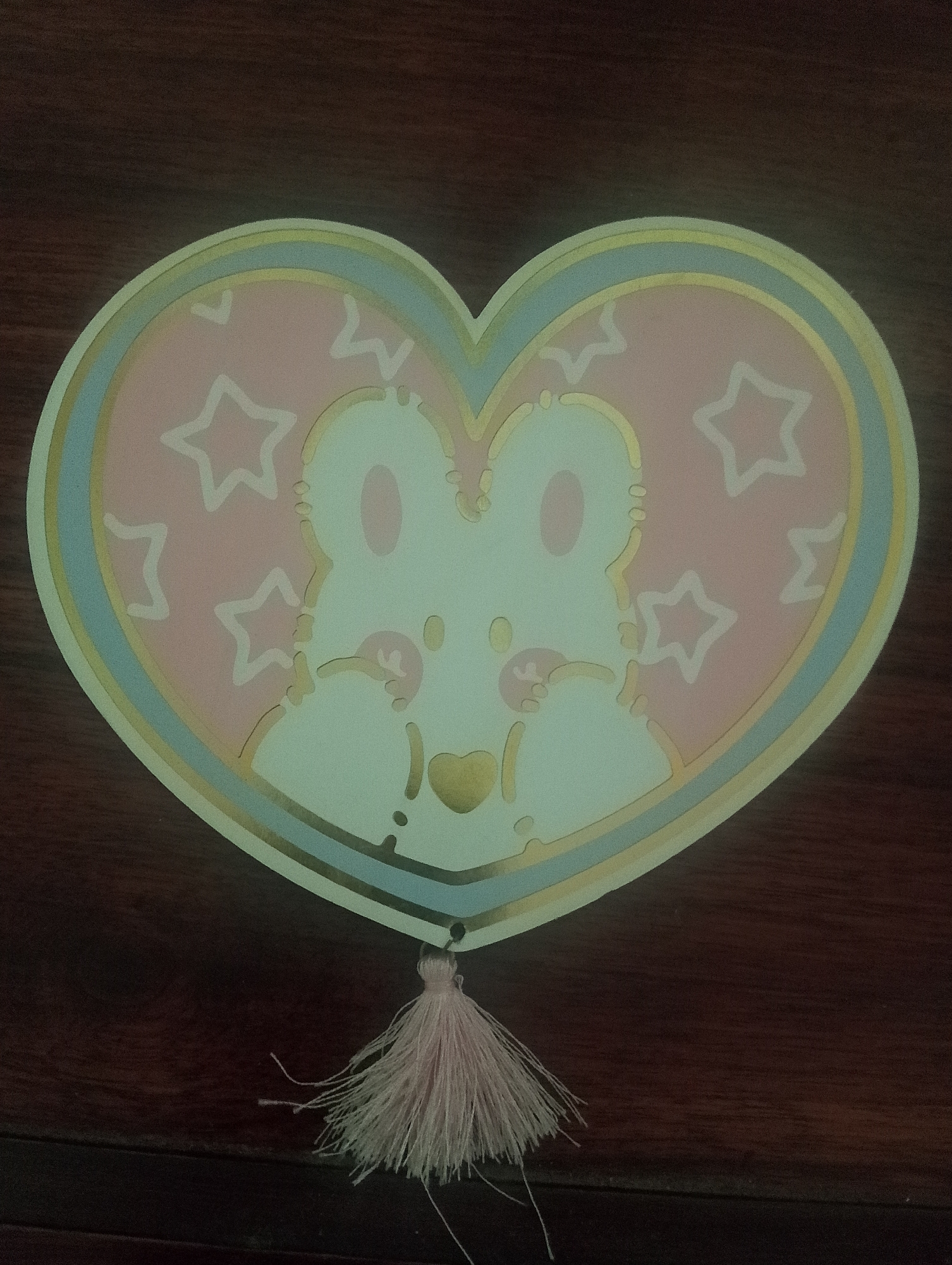 A heart-shaped object with a pastel pink background, featuring a white cartoon bear character with pink accents. The bear has large oval ears, two oval eyes, a small gold nose, and paw-like hands. The background is adorned with white stars and zig-zag shapes. The object has a light blue and gold border. A light pink tassel hangs from the bottom center of the object. The object is placed on a dark wooden surface, likely a table, with visible wood grain. The image was captured indoors. The location of Serbelawan, Indonesia is not discernible from the image.