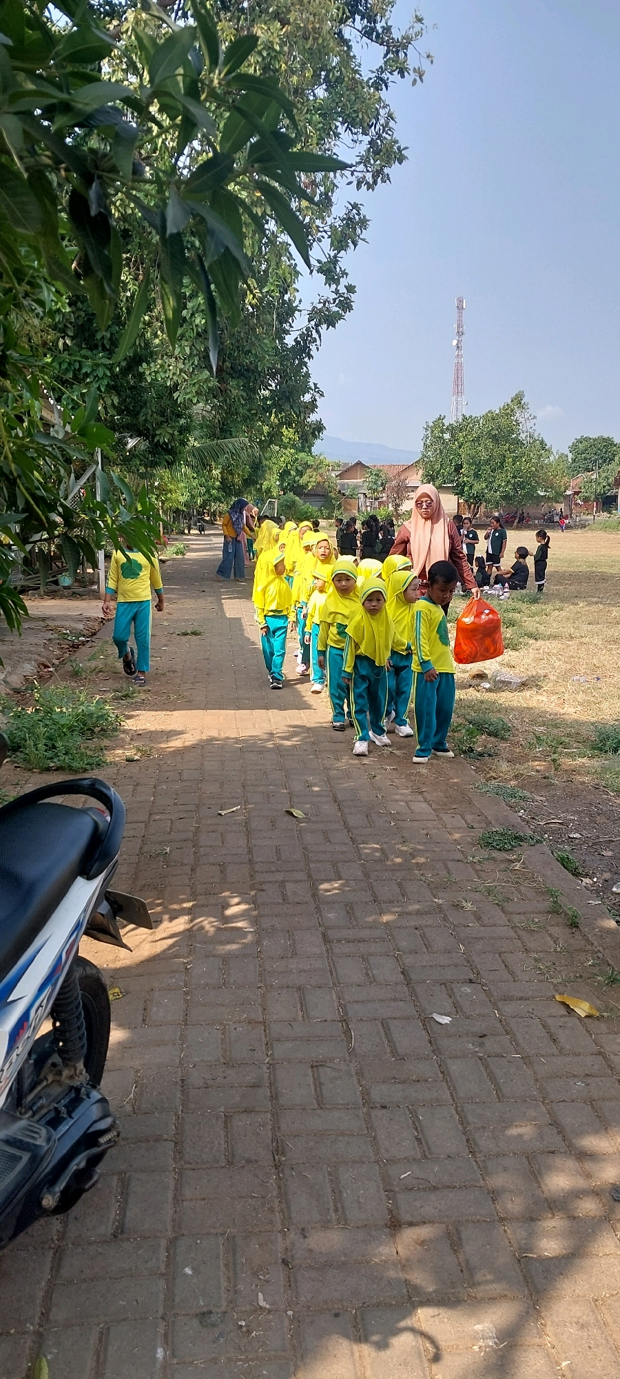 In the city of Keling, Indonesia, a group of young children dressed in matching yellow tops and teal pants, some wearing yellow headscarves, are walking in a line along a brick pathway. The children appear to be of preschool or early elementary age. Behind the children, an adult woman wearing a peach-colored headscarf and a brown top is walking, holding a large orange plastic bag. Several other children and adults are visible in the background, some sitting on the ground and others standing. The environment includes trees with green foliage casting shadows on the pathway, a distant cell tower, and residential buildings. A motorcycle is parked on the left side of the pathway in the foreground. The scene suggests an outdoor activity or field trip.
