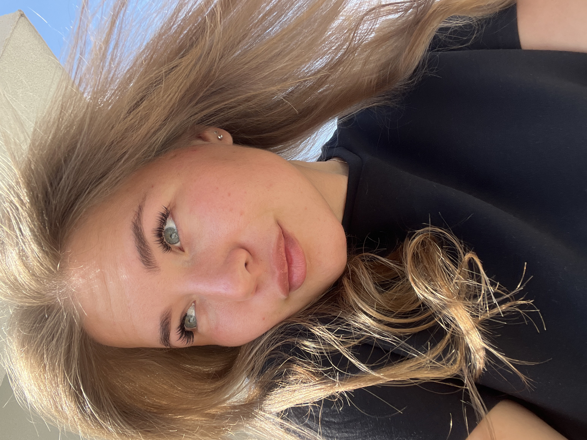 The image displays a close-up portrait of a young woman, captured from a low, upward-looking angle. Her head is tilted, giving the photograph a casual, possibly selfie-like quality.
The woman has long, straight to slightly wavy hair that appears to be a light brown or blonde color, with noticeable highlights from the bright sunlight. Her hair cascades around her face and shoulders, appearing somewhat windswept.
Her facial features are prominent. She has fair skin, with a natural, unblemished complexion, though some slight redness is visible on her cheeks. Her eyes are a striking light color, possibly blue or green, and she has long, dark eyelashes, which appear well-defined and curled. Her eyebrows are neatly shaped. Her nose is small, and her lips are full with a natural, light pink hue, slightly parted. A subtle, clear stud earring is visible in her right earlobe.
She is wearing a simple, plain black top or shirt, the fabric appearing soft and possibly cotton.
The lighting in the image is bright and natural, suggesting an outdoor setting on a sunny day. The sunlight illuminates her hair and face, creating highlights. In the upper left background, a clear, bright blue sky is visible, along with a partial view of a light-colored, possibly white or cream-colored, structure or object, which could be part of a building or an awning.
The woman looks directly into the camera with a soft, relaxed expression, verging on a faint smile. There is no visible text in the image.