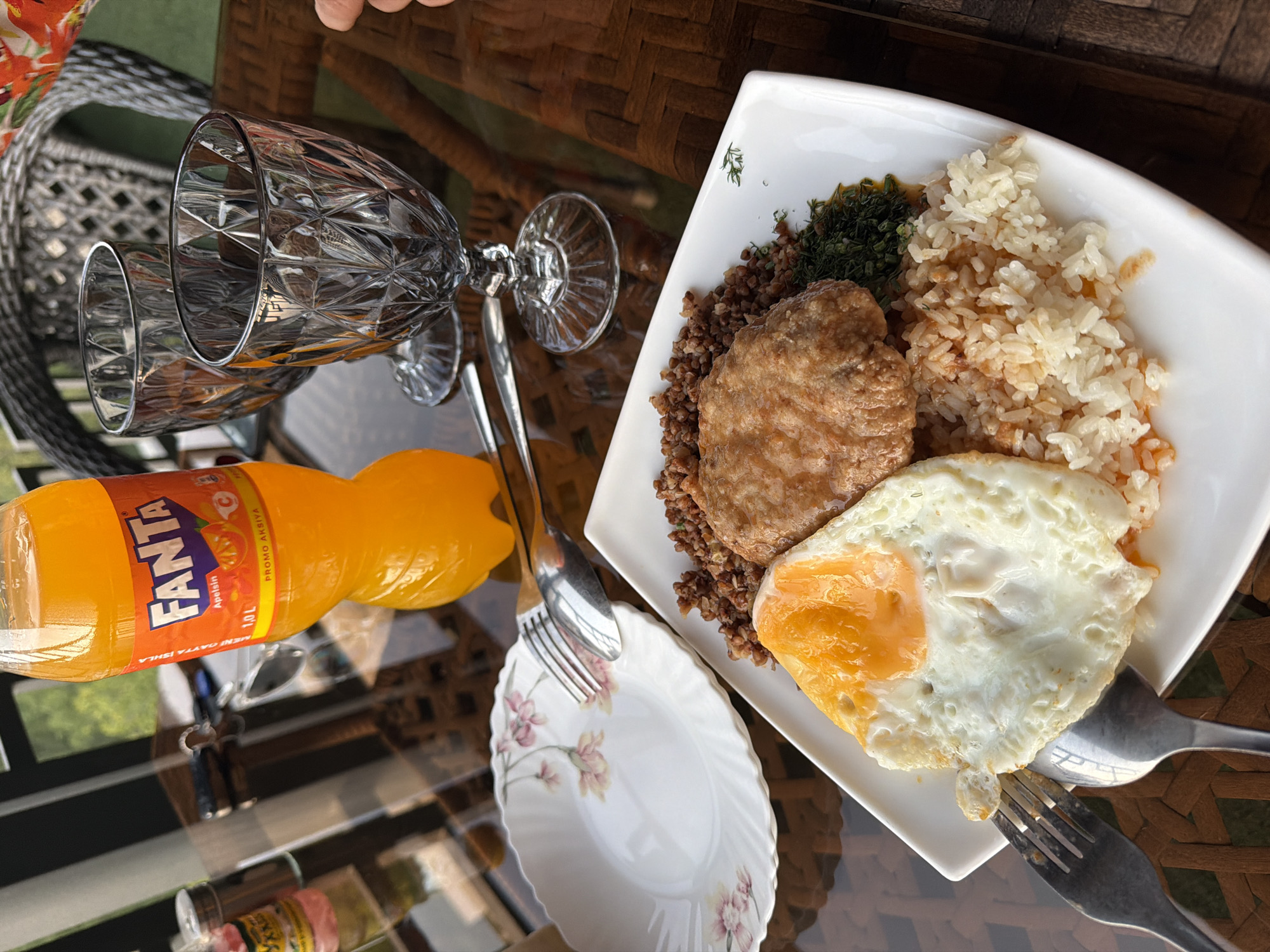 A top-down, eye-level, medium shot captures a dining scene on a reflective, dark brown woven table, possibly in the city of Nanay, Uzbekistan, given the context. The focus is on a white rectangular plate filled with a meal. The meal consists of a sunny-side-up egg with a bright orange yolk, a portion of fluffy white rice with some red sauce, and a pile of dark brown buckwheat groats topped with a golden-brown, oval-shaped cutlet. A small cluster of chopped green herbs is visible on the right side of the plate.
To the left of the plate, a delicate white floral-patterned plate with pink and yellow flowers is positioned, holding a fork and spoon. In the background, an orange Fanta bottle with a label indicating "1.0 L" stands upright. Two clear, cut-glass goblets, reflecting the light, are placed behind the Fanta bottle. Further in the background, a portion of a dark gray woven chair is visible. The lighting suggests an outdoor setting, possibly during daylight.