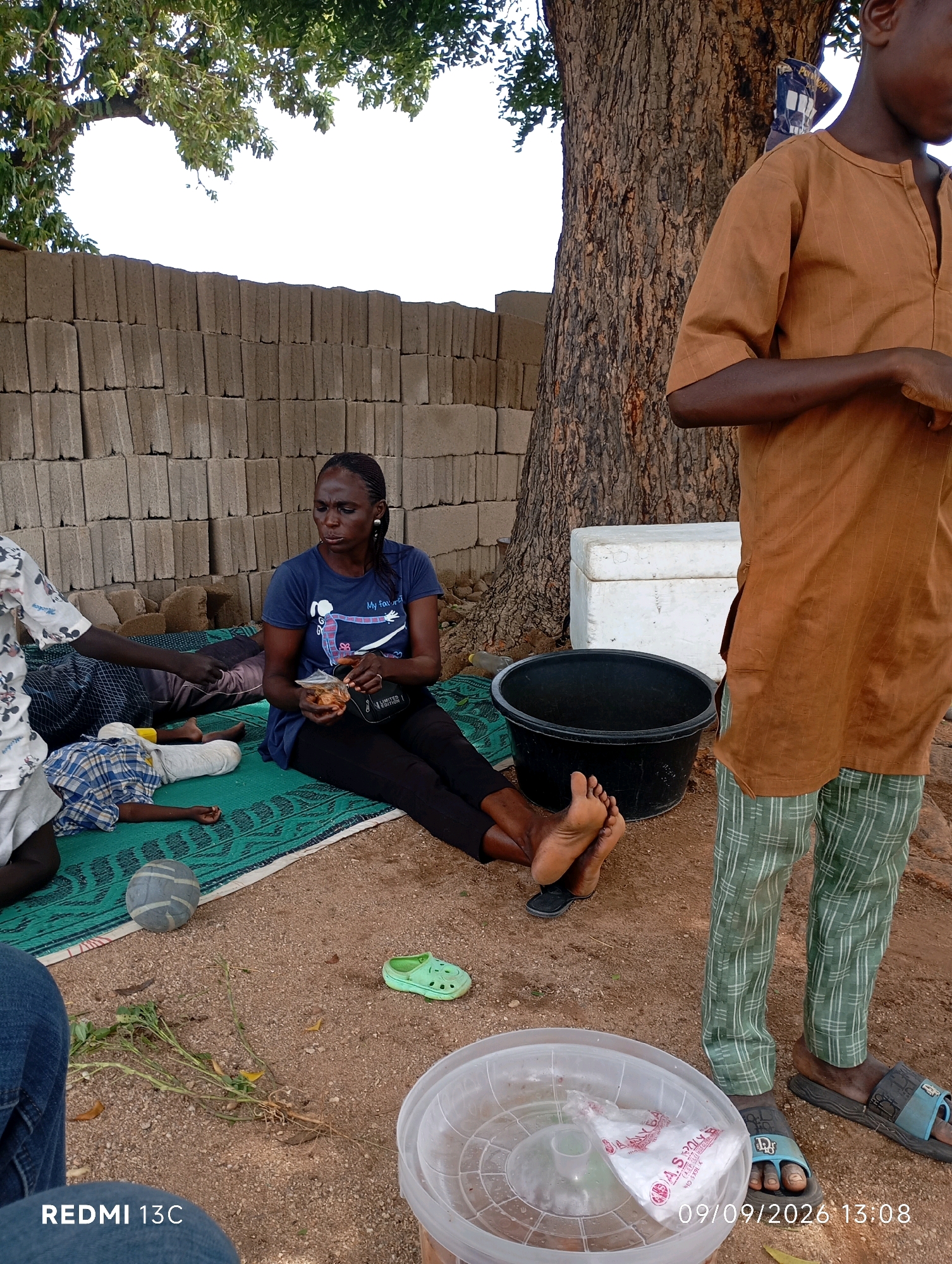 In Shendam, Nigeria, a woman sits on a green mat in front of a stack of concrete blocks, holding a small bag of food. She wears a dark blue t-shirt and black pants. To her left, a child lies on the mat, wrapped in a blue and white checkered cloth. Another person, partially visible on the far left, wears a white shirt with a Mickey Mouse print. A large black bucket and a white cooler are positioned behind the woman and child. A young person in a brown tunic and green checkered pants stands to the right, facing away from the camera. The ground is covered in dirt and scattered debris. In the foreground, a clear plastic container with a white plastic bag inside is visible, along with a green child's shoe and a blue and grey ball. The scene is set outdoors, with a large tree providing shade. The timestamp on the image indicates 09/09/2026 at 13:08.