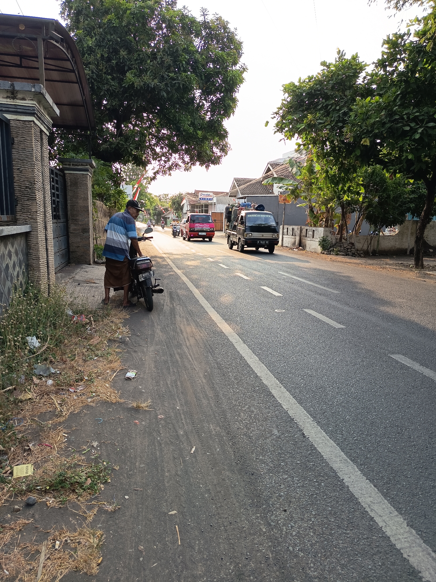 The image depicts a street scene in Kandangjati Wetan, Indonesia, under a clear sky with soft lighting, suggesting it might be late afternoon. The focus is on a man standing next to a motorcycle on the side of a two-lane asphalt road. The man, wearing a striped blue and white shirt and a brown sarong, is looking towards the street. The motorcycle is black and parked on the shoulder of the road.
The roadside is somewhat unkempt, with dry grass, weeds, and scattered litter visible. To the left of the man, a stone wall and a metal gate suggest a property entrance. Beyond the wall, a building with a covered entrance is visible.
The road itself has white lane markings. In the distance, several vehicles are traveling: a red car, a dark colored small truck, and two motorcycles. The truck appears to be carrying some items in its bed. The environment is characterized by trees lining the road, with houses and buildings visible in the background. The overall impression is a typical street scene in a residential or semi-commercial area of an Indonesian town.
