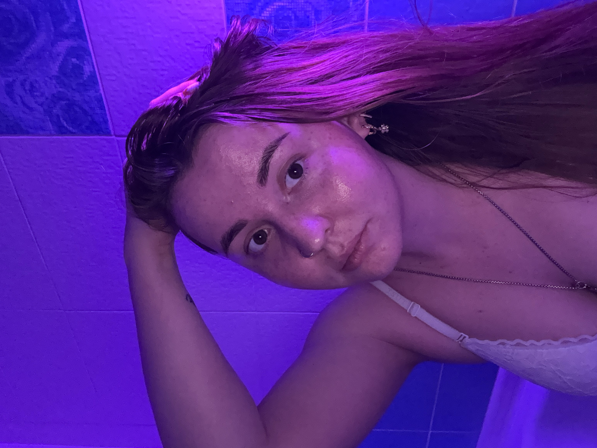 A young woman is prominently featured in a close-up, angled portrait. Her head is tilted, and she looks directly at the camera with a direct gaze. Her long hair, a mix of brown with distinctive purple or pink highlights, frames her face. Her skin appears luminous or moist, possibly from sweat or the effects of the lighting. She has a subtle nose piercing and a delicate chain necklace around her neck. An earring, featuring multiple small decorative elements, is visible on her right ear. She wears a white bra or bralette, the lace detail of which is visible on the strap and cup. Her left arm is bent, with her hand positioned near her head, suggesting a deliberate pose. The background consists of tiled walls; the upper section displays blue tiles, while the overall scene is immersed in a vibrant, artificial purple light, creating an atmospheric, perhaps club-like or intimate indoor setting, potentially a bathroom. The intense colored lighting makes the time of day indistinguishable. No text is visible. The location context is Izhevsk, Russia.