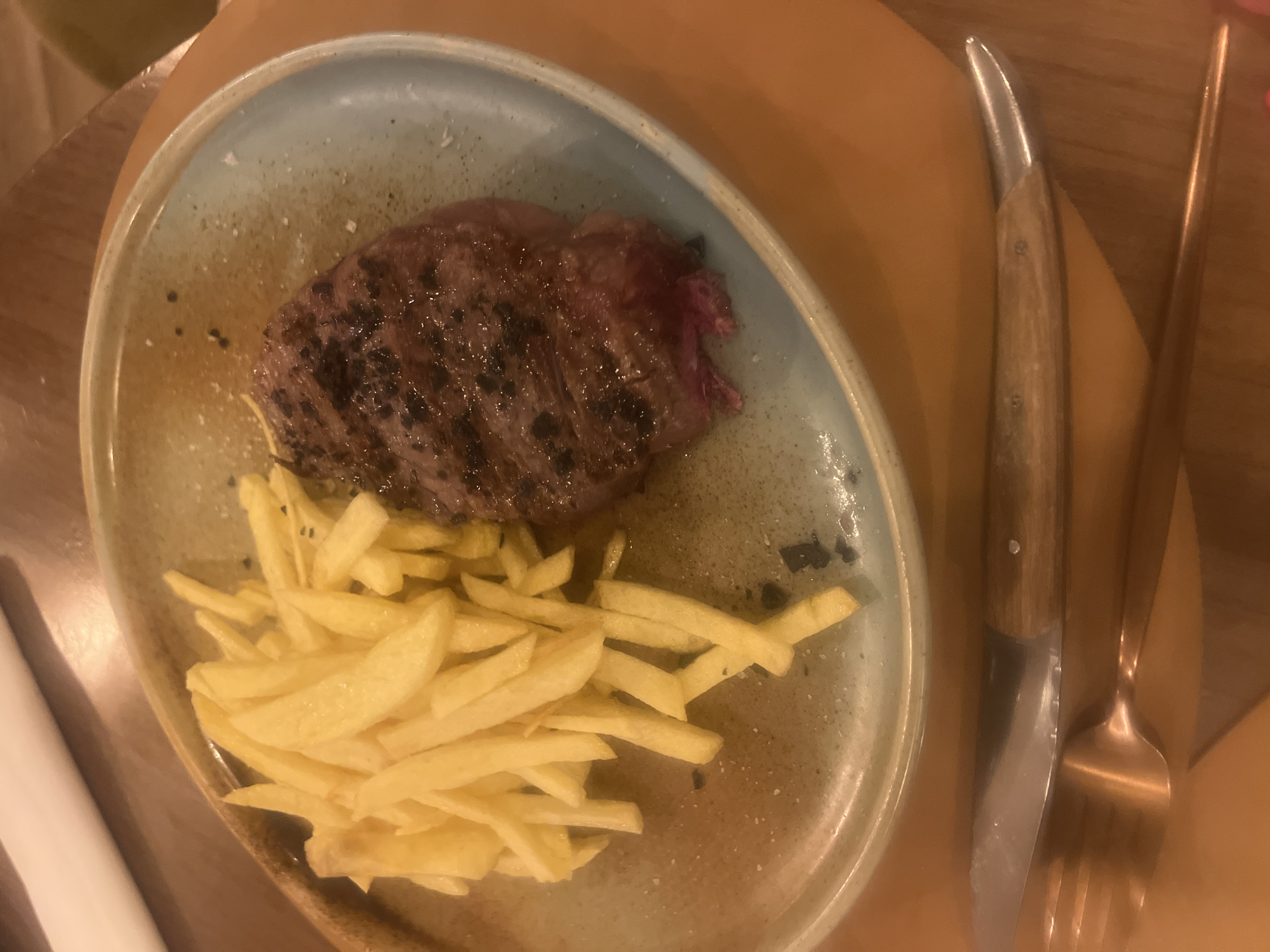 A plate of steak and fries sits on a table. The steak, cooked to medium-rare, is slightly charred on the outside. The fries are pale yellow and appear to be lightly salted. A knife with a wooden handle and a fork are positioned to the right of the plate. The plate is a light blue-grey color with a speckled texture. The table appears to be made of wood. The location cannot be confirmed as Sant Pere I Sant Pau, Spain, based on the image alone. There is no information regarding people, actions, or interactions in the image.