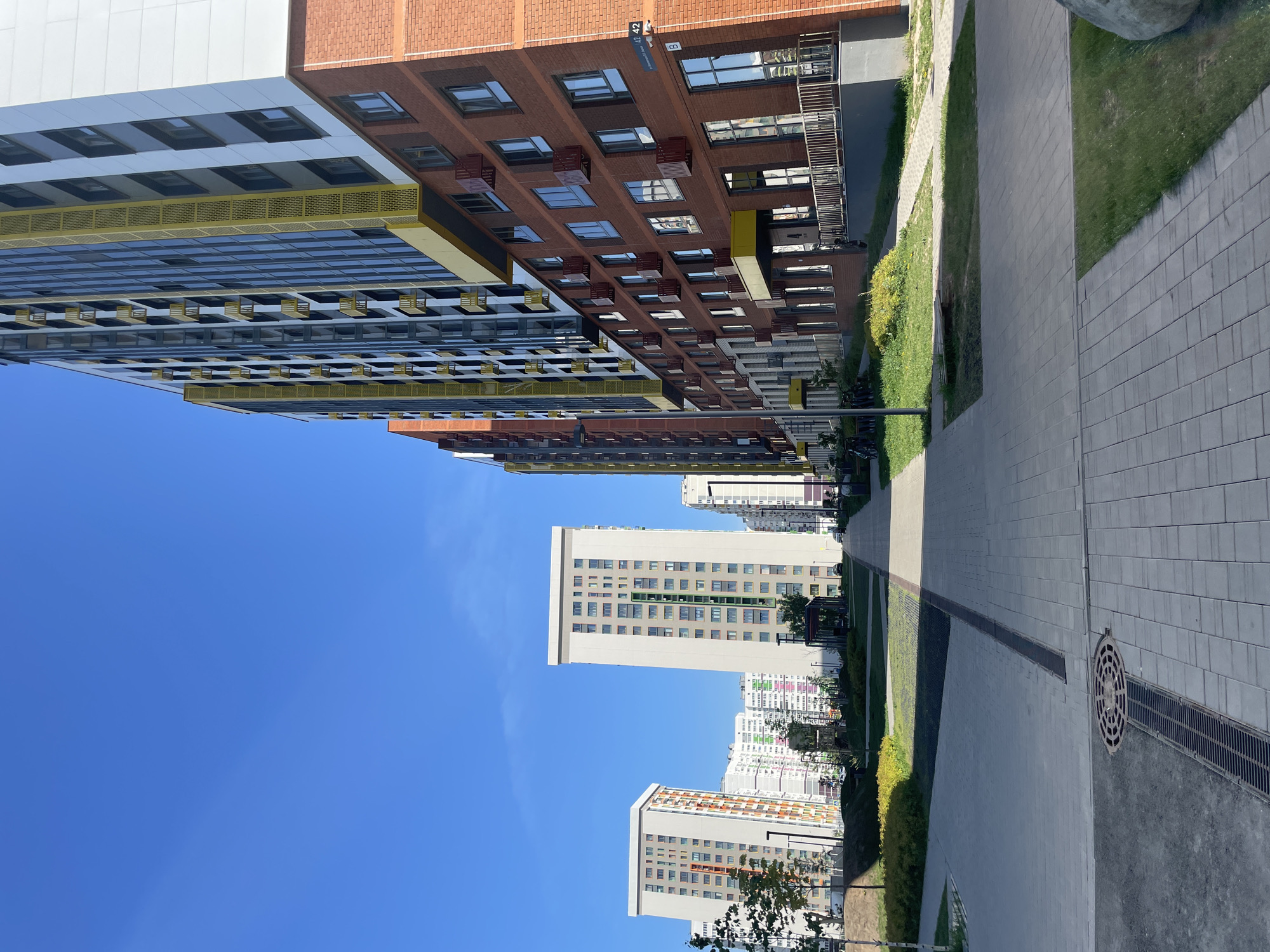 The image depicts a streetscape in Izhevsk, Russia. Multiple modern apartment buildings are visible, varying in height and architectural style. The buildings are constructed with different materials and feature colorful facades, including brick, white panels, and yellow accents. A pedestrian walkway runs along the right side of the frame, composed of light gray paving stones and lined with narrow strips of green grass. A dark, linear drainage grate runs along the walkway. The sky is a clear, bright blue. There are no people visible in the image.