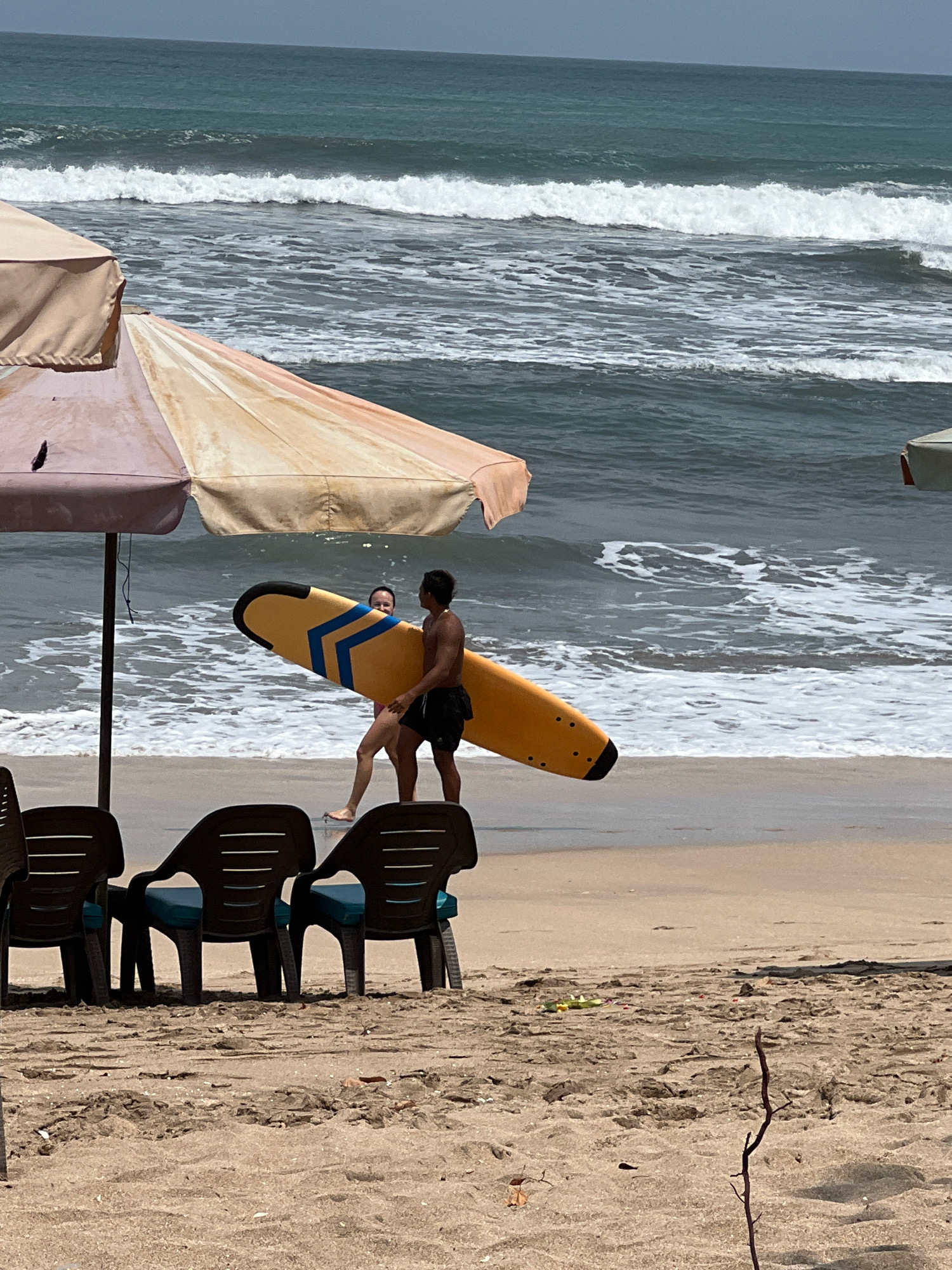 In Kuta, Indonesia, a couple walks across the sandy shore with a yellow surfboard. The man, wearing black shorts, carries the board under his arm. The woman, in pink shorts, walks beside him, looking towards the camera. Two beach umbrellas provide shade, one with a faded pink and beige canopy, the other partially visible and light brown. In the foreground, three empty dark brown plastic beach chairs with teal cushions are arranged on the sand. The ocean in the background features moderate waves with white foam, and the sky is overcast.