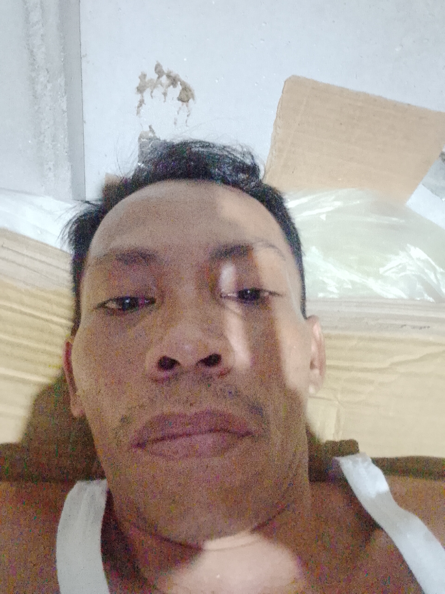 A close-up selfie depicts a middle-aged man wearing a white tank top. The photo appears to be taken indoors, with a ceiling visible in the background. There's some peeling paint and debris on the ceiling. Behind the man are items including cardboard. The photo's location is Bukit Ketok, Indonesia.
