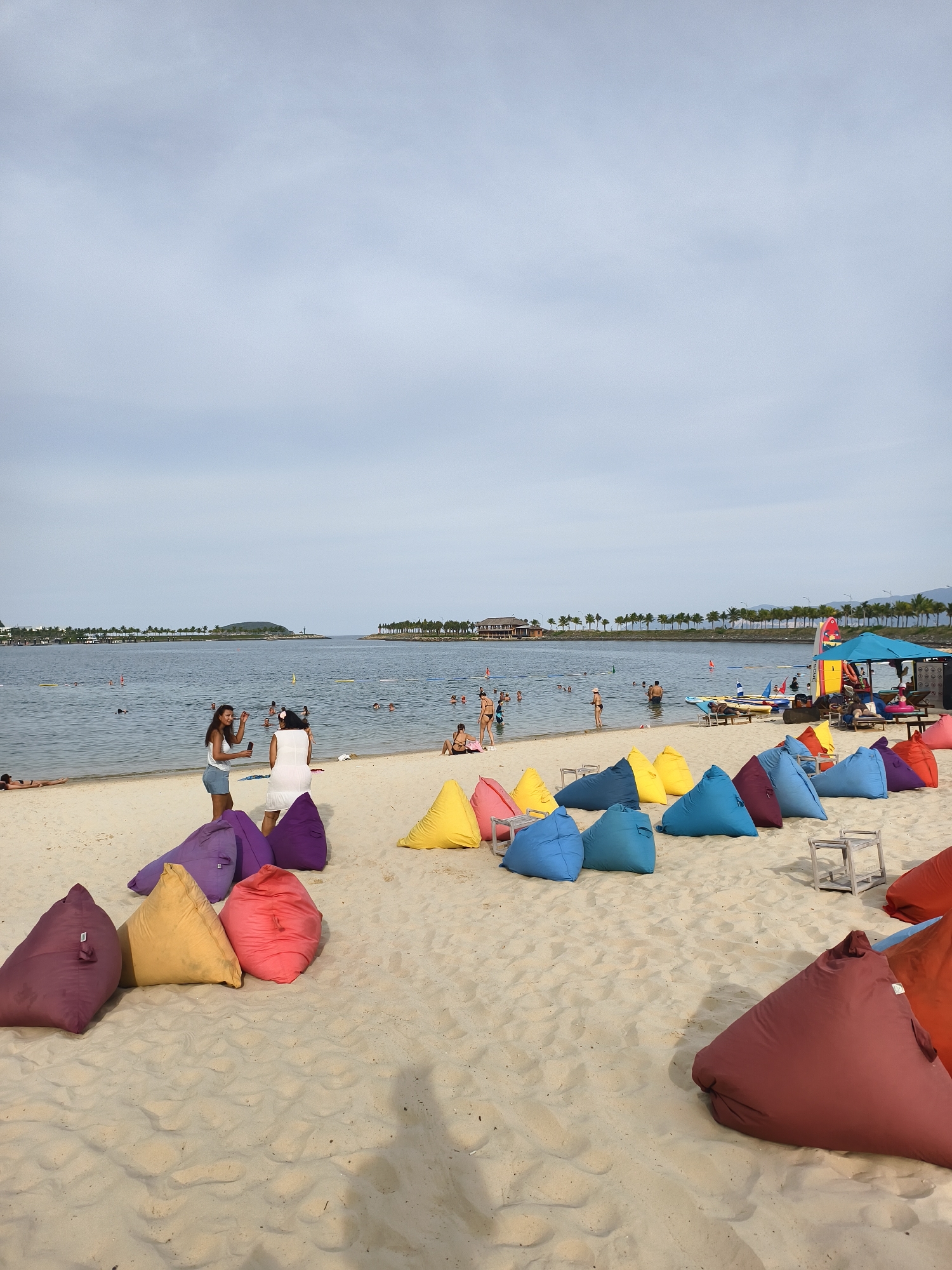 This image depicts a beach scene in Bac Nha Trang Ward, Vietnam. The foreground is dominated by a row of colorful beanbag chairs, in shades of purple, yellow, red, and blue, resting on the sand. Several small, metallic chairs are also visible among the beanbags. In the middle ground, two women are standing on the sand near the water's edge, one in a white dress and the other in a white top and denim shorts. The woman in white appears to be taking a photo with her phone. A few other individuals are sunbathing on the sand behind them. The background features a calm body of water, with several people swimming and wading. A distant coastline is lined with palm trees and some buildings. The sky is overcast, with a light haze.