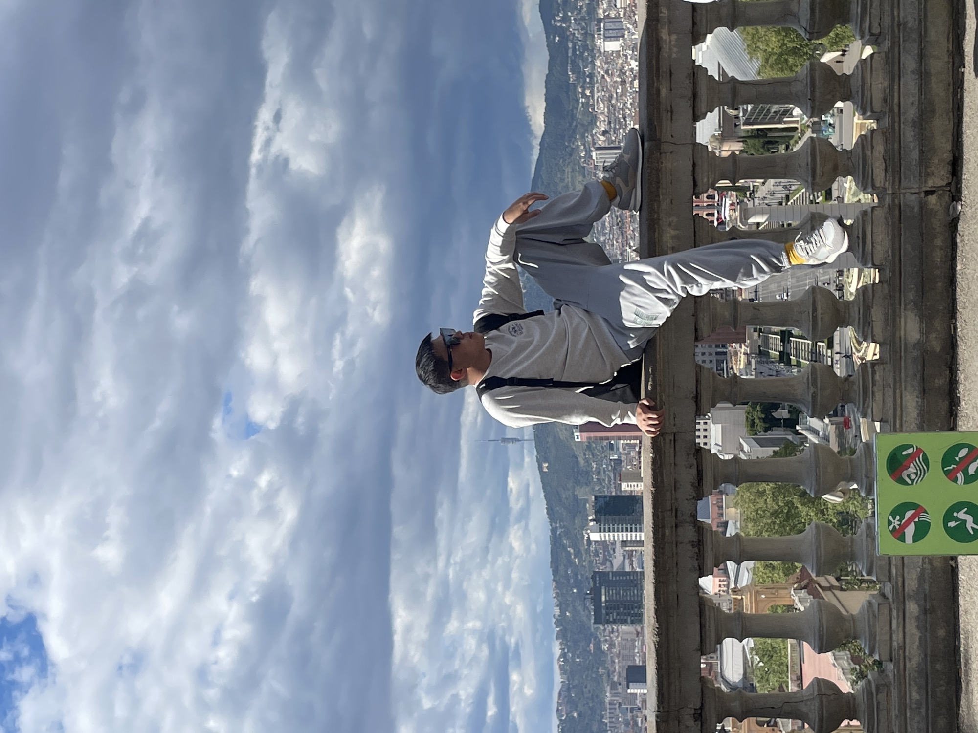 A man is sitting on a ledge overlooking a city in Spain. He is wearing a light gray sweatshirt, gray sweatpants, sunglasses, and sneakers. His left knee is raised, and he is resting one arm on it. The city behind him appears dense, with tall buildings. There is a mountain in the background. The sky is partly cloudy. A sign can be seen to his right that indicates swimming, falling and diving is not permitted. The latticed stone border suggests a lookout point. It is likely this photograph was taken in Barcelona, Spain.