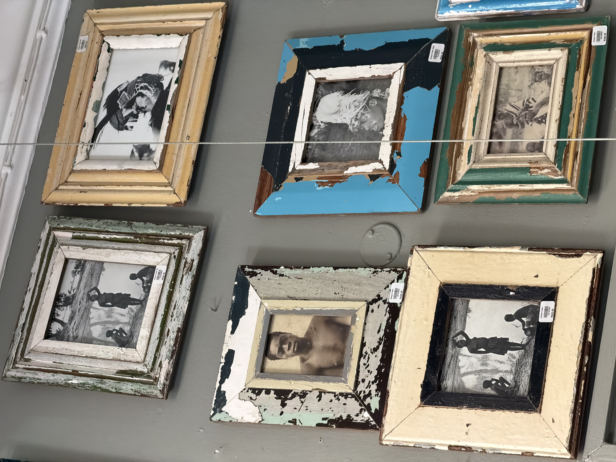 The image displays six framed, black and white photographs arranged on a plain gray wall, suggesting an interior retail or gallery setting. All frames are intentionally distressed, featuring peeling paint in various colors such as cream, blue, green, and black, giving them a rustic or antique aesthetic. A thin wire runs horizontally across the middle of the display.
The photographs predominantly feature people in what appear to be historical or cultural contexts:
1. **Top Left:** A cream-colored frame holds a dynamic shot of a person, possibly a woman, with long hair in motion. A price tag on the upper left corner reads "R300.00".
2. **Top Middle:** A blue and black distressed frame encircles a close-up portrait of a person's face with wild, flowing hair. A partially obscured price tag is on the right.
3. **Top Right:** A green and cream frame contains an image of two individuals, possibly children, wearing striped tops in an outdoor setting. A visible price tag reads "R440.00".
4. **Bottom Left:** A green and white distressed frame showcases three figures – two adults and a child – engaged in an activity by a body of water. One adult is standing, possibly carrying something, while another is stooping, and a child is seated. A price tag on the top right reads "R400.00".
5. **Bottom Middle:** A multi-colored distressed frame (white, black, blue, wood) holds a portrait of a shirtless man looking directly at the viewer with a neutral expression. A price tag is visible on the upper right, but the text is largely unreadable.
6. **Bottom Right:** A cream and black frame displays a scene similar to the bottom left, featuring three figures by water, with one adult standing and seemingly carrying something on their head, another stooping, and a child seated. The photo is rotated, making the figures appear to stand on the right side of the frame. A price tag reads "R400.00".
The consistent black and white aesthetic and the "R" currency symbol on the visible price tags (Rand) align with the provided context of Cape Town, South Africa, suggesting these are antique or vintage-style photos for sale. The overall impression is one of curated, vintage-inspired decor.