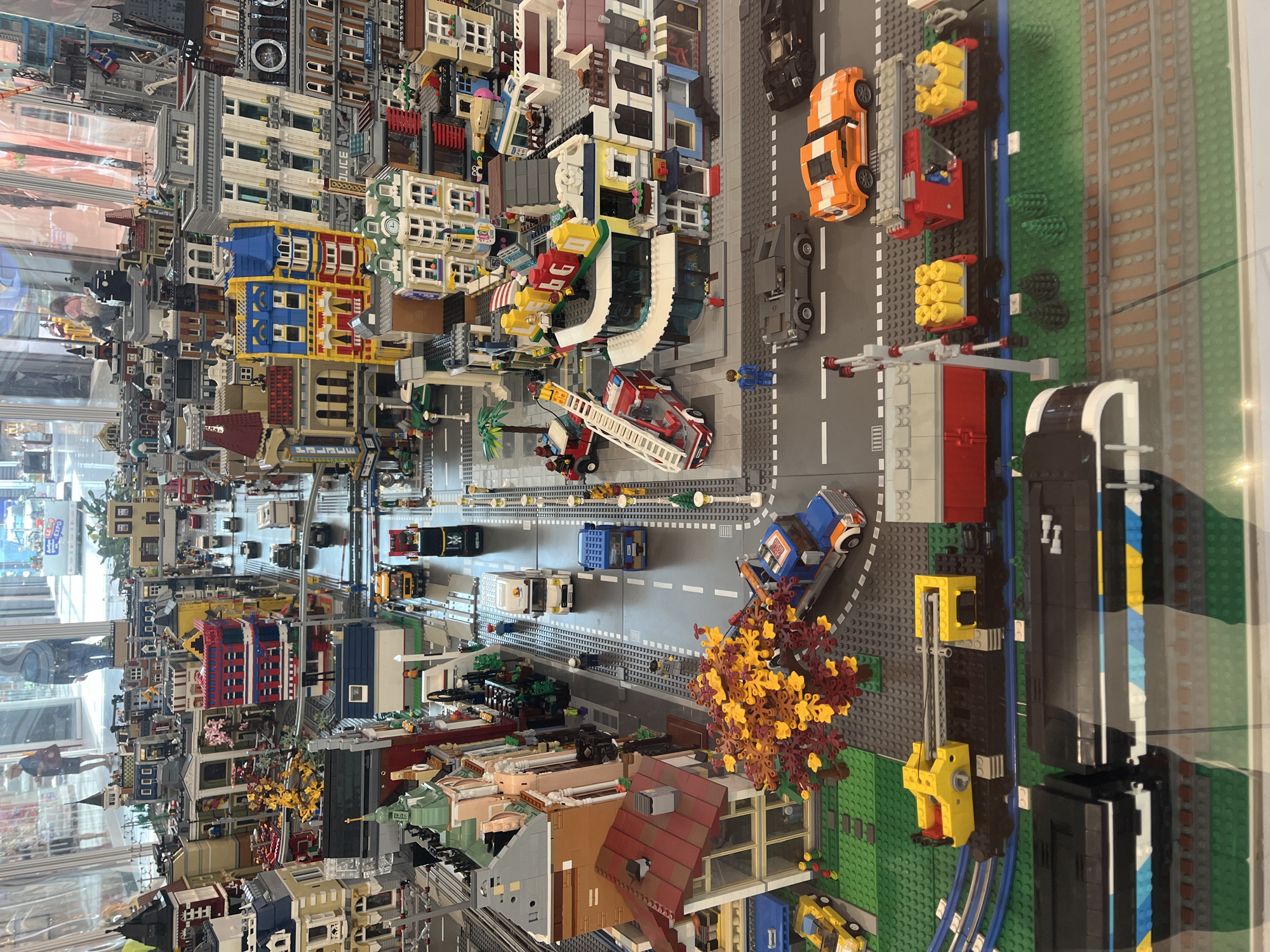 This is an overhead, slightly angled shot of a large, detailed Lego city diorama. The diorama depicts a bustling urban environment with various buildings, vehicles, and figures. The buildings are constructed from a variety of Lego bricks and range in architectural style, including what appear to be residential apartments, commercial storefronts, and public buildings.
Vehicles are scattered throughout the scene, including cars, trucks, a fire engine with an extended ladder, and a train with multiple carriages. Some of the vehicles appear to be in motion or stopped at intersections. Lego figures are present, some standing on sidewalks or near buildings, and others appearing to be driving the vehicles or working on the train.
The environment includes roads, sidewalks, green spaces with Lego trees, and what appears to be a park area with a Lego tree in autumnal colors. The diorama is set against a blurred background of what looks like a public exhibition space, with reflections visible on a glass barrier in the foreground. The overall impression is a meticulously crafted and detailed miniature city.