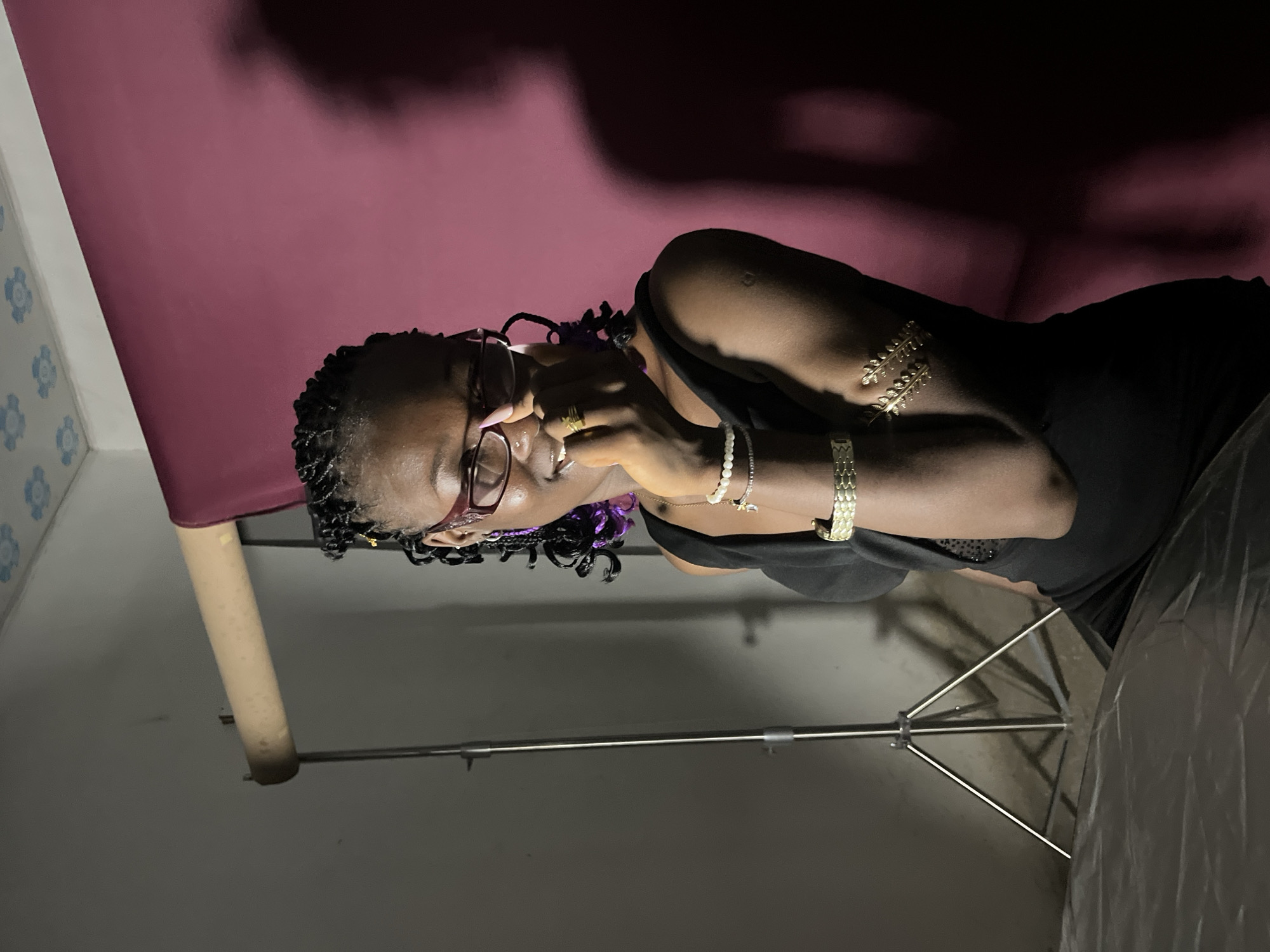 A medium shot captures a young Black woman in a dimly lit room, possibly a studio, given the presence of a light stand and a backdrop. The woman is seated, and her face is partially obscured as she brings her right hand to her mouth, fingers touching her lips. She is wearing glasses with reddish frames and has dark braided hair with strands of purple woven into it. She is adorned with several pieces of jewelry, including bracelets on both wrists and upper arms, and rings on her fingers. Her attire is a black sleeveless top. The background consists of a plain light-colored wall on the left and a deep pink backdrop on the right, casting a shadow of the woman onto it. The ceiling has a decorative pattern. The floor in the foreground is covered with a grey, translucent material. The overall lighting creates a moody atmosphere with strong contrasts and shadows.