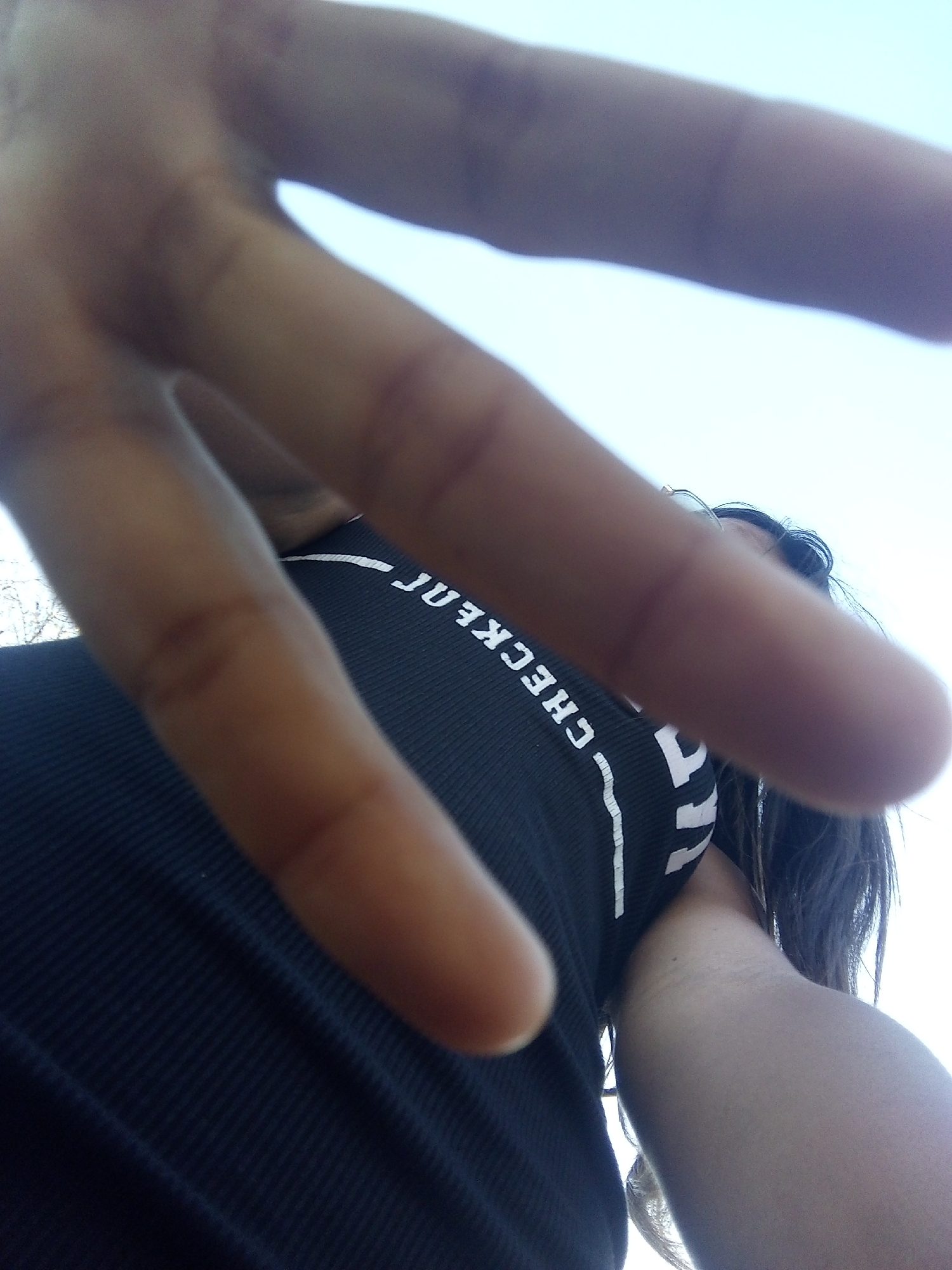 A close-up, low-angle shot shows a person with a hand extended towards the camera. The person is wearing a black ribbed top with the word "CHECKER" printed vertically in white letters. Strands of dark hair are visible near the right side of the frame. The background shows a pale blue sky. The location in Juan Griego, Venezuela is not identifiable from the image.