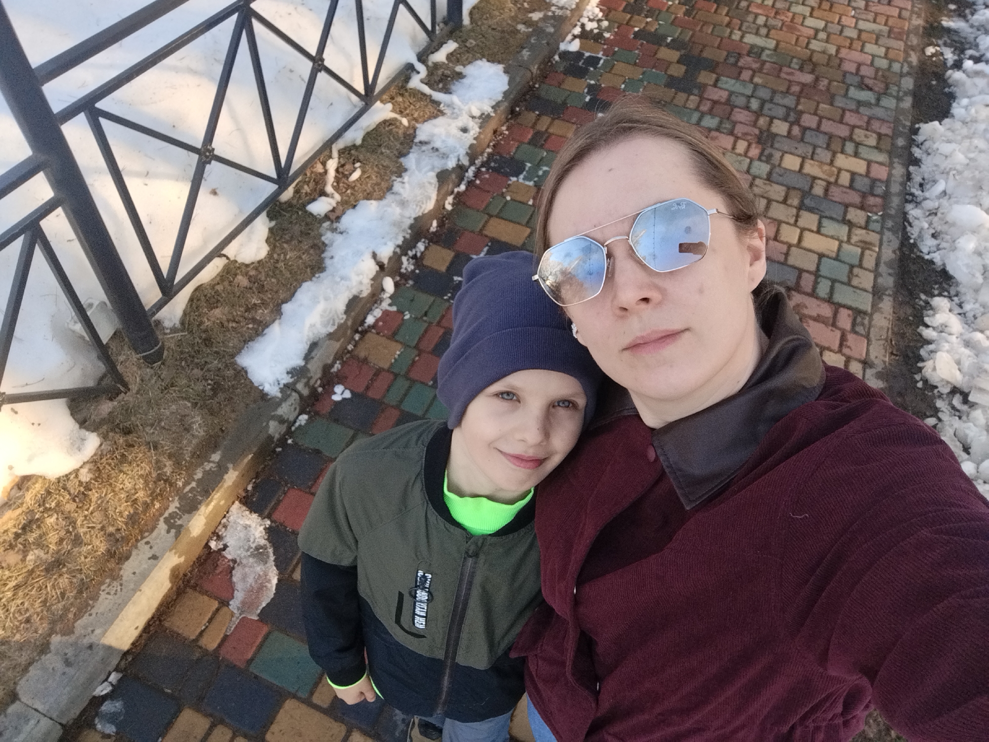 A medium, high-angle shot depicts a woman and a child standing closely together outdoors. The woman, positioned on the right side of the frame, wears reflective sunglasses and a dark red, corduroy-like jacket with a dark-colored collar. The child, standing on the left and slightly behind the woman, wears a dark-colored beanie and a jacket with a green interior.
The environment consists of a paved walkway with colorful bricks. To the left of the walkway is a black metal railing, beyond which lies snow and grass. Patches of snow are also visible along the edge of the walkway.
Based on visual analysis, the location is most likely in the city of Izhevsk, Russia.