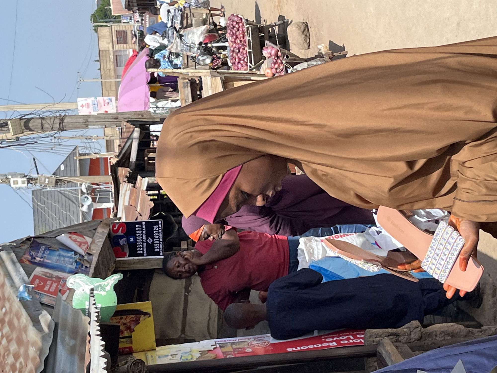 The image shows a scene in a busy, presumably outdoor market or commercial area, potentially in Jos, Nigeria, based on the likely African ethnicity of the individuals and the sign with a local Nigerian phone number.
In the foreground, there's an adult wearing a brown hijab-like head covering. The person is holding a pair of light pink sandals embellished with decorative stones or beads. Below, a person wearing a maroon shirt rests their head on their hand.
In the background, the scene reveals a cluttered urban environment with various shops or stalls. There are signs, electrical wires, and a collection of goods. A sign is visible with a telephone number: “8233603877 BASIRU ABDULLAHI” and "9550749726 BASIRU ABDULLANI". There are metal and corrugated iron structures visible, with other merchandise and items possibly for sale, including onions.