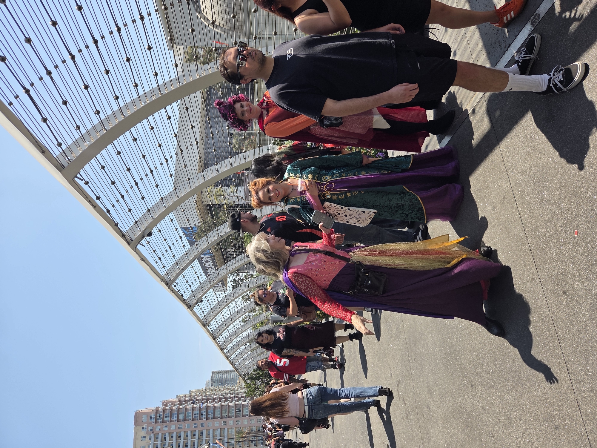 An outdoor, daytime image shows a group of people walking on a sidewalk, with a modern, curved pedestrian overpass in the background. The overpass is constructed of metal beams and translucent panels, with a grid of wires and small spherical fasteners forming its sides. In the distance, tall, light-colored apartment buildings are visible, as is a building with a sign that reads "BEACH." A few people in the foreground are dressed in costumes. To the left, a woman with long brown hair wears a light pink crop top and blue jeans. To her right, a man in a red shirt with the number 5 on it walks by. In the center, two women are dressed as characters from Hocus Pocus. One wears a green and purple gown, and the other is in a reddish-orange outfit with a ruffled collar and a pink wig. Another man in a black t-shirt and shorts stands to the right of the costumed women, facing forward. The ground is paved with concrete, and the sky is clear and blue. The scene appears to be in Long Beach, California, given the architectural style of the buildings and the pedestrian overpass, which resembles structures found in that area.
