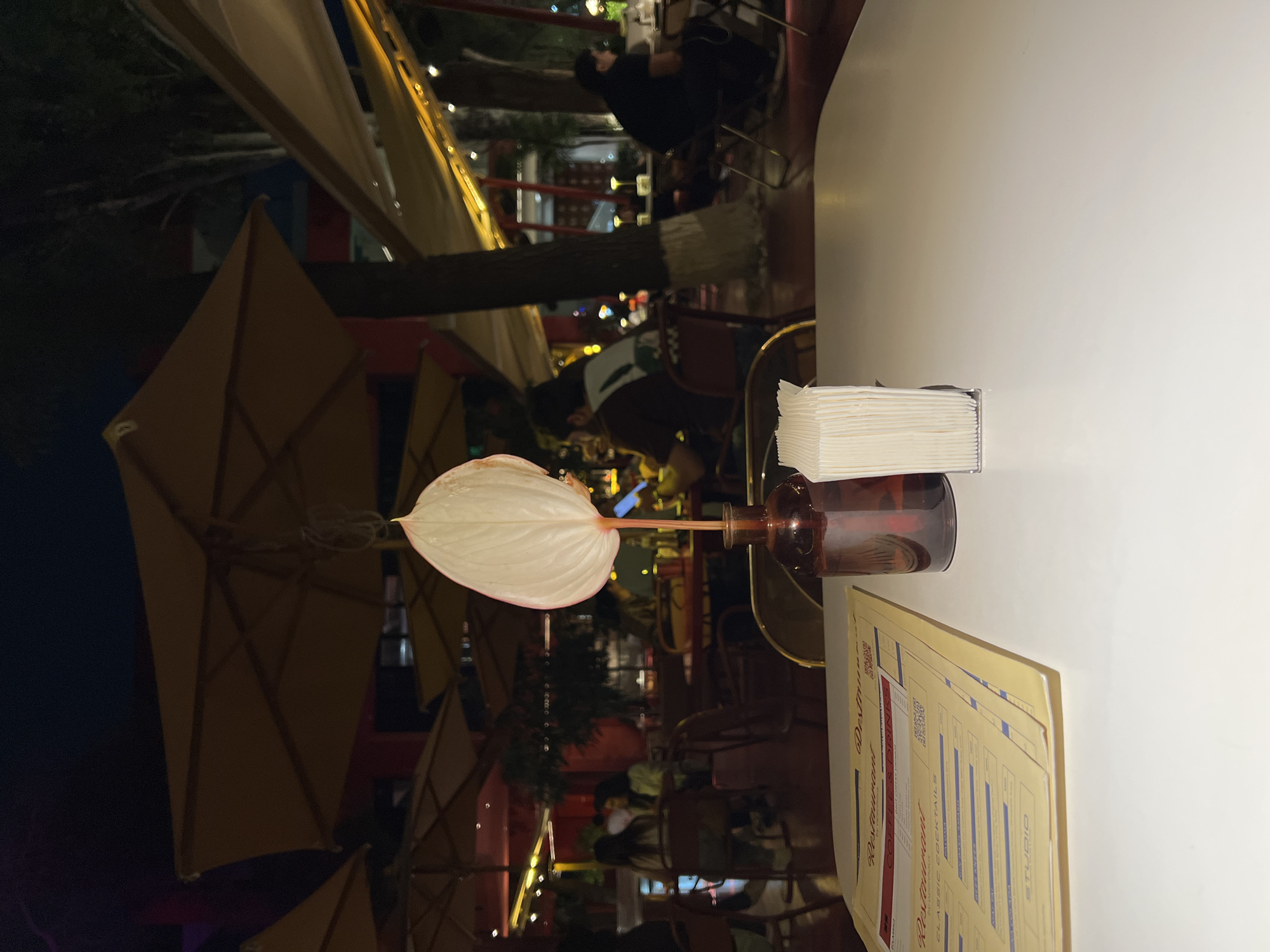 An outdoor seating area of a restaurant in Yerevan, Armenia is depicted in this nighttime photograph. The foreground is dominated by a white table, on which rests a menu, a bottle with a single white flower, and a stack of napkins. The menu, yellowed with age, displays sections for "Restaurant," "Coffee & Drinks," and "Classic Cocktails." In the background, patrons are seated at tables under large, amber-colored umbrellas and a beige canopy. Trees with visible bark patterns are interspersed among the seating areas, and string lights provide illumination. The overall atmosphere is dimly lit, suggesting evening hours.
