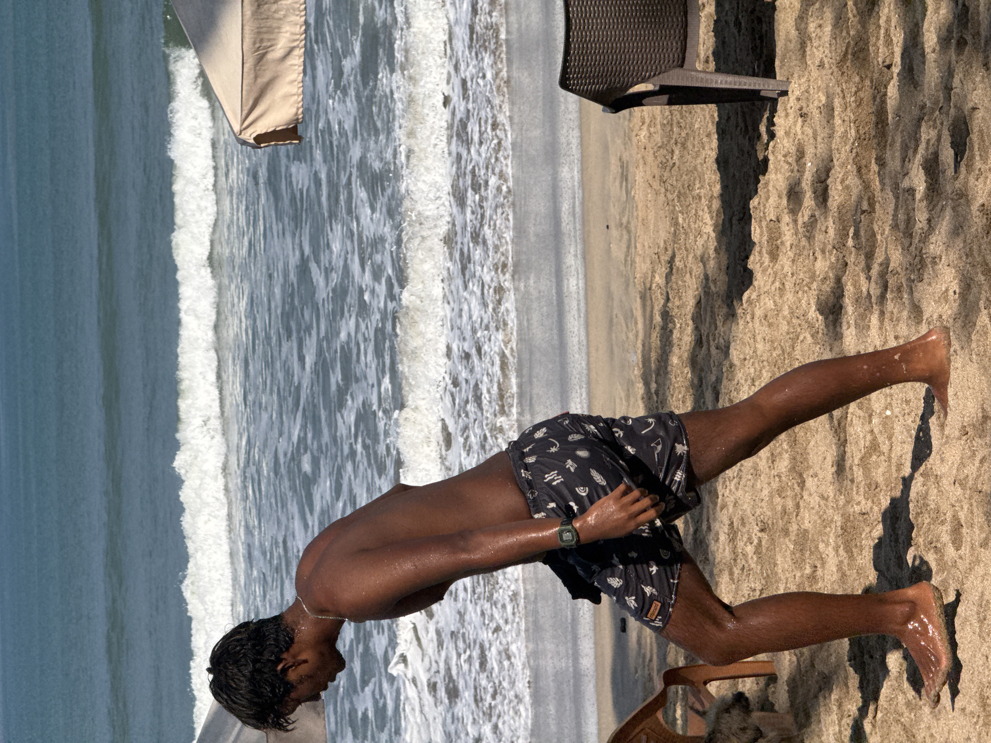 A young man walks across a sandy beach in Kuta, Indonesia. He is wearing dark swim trunks with a white pattern and a silver chain. His skin is wet and glistening in the sun. Behind him, the ocean waves crash onto the shore. Beach umbrellas and chairs are visible in the background. The sun casts long shadows on the sand.