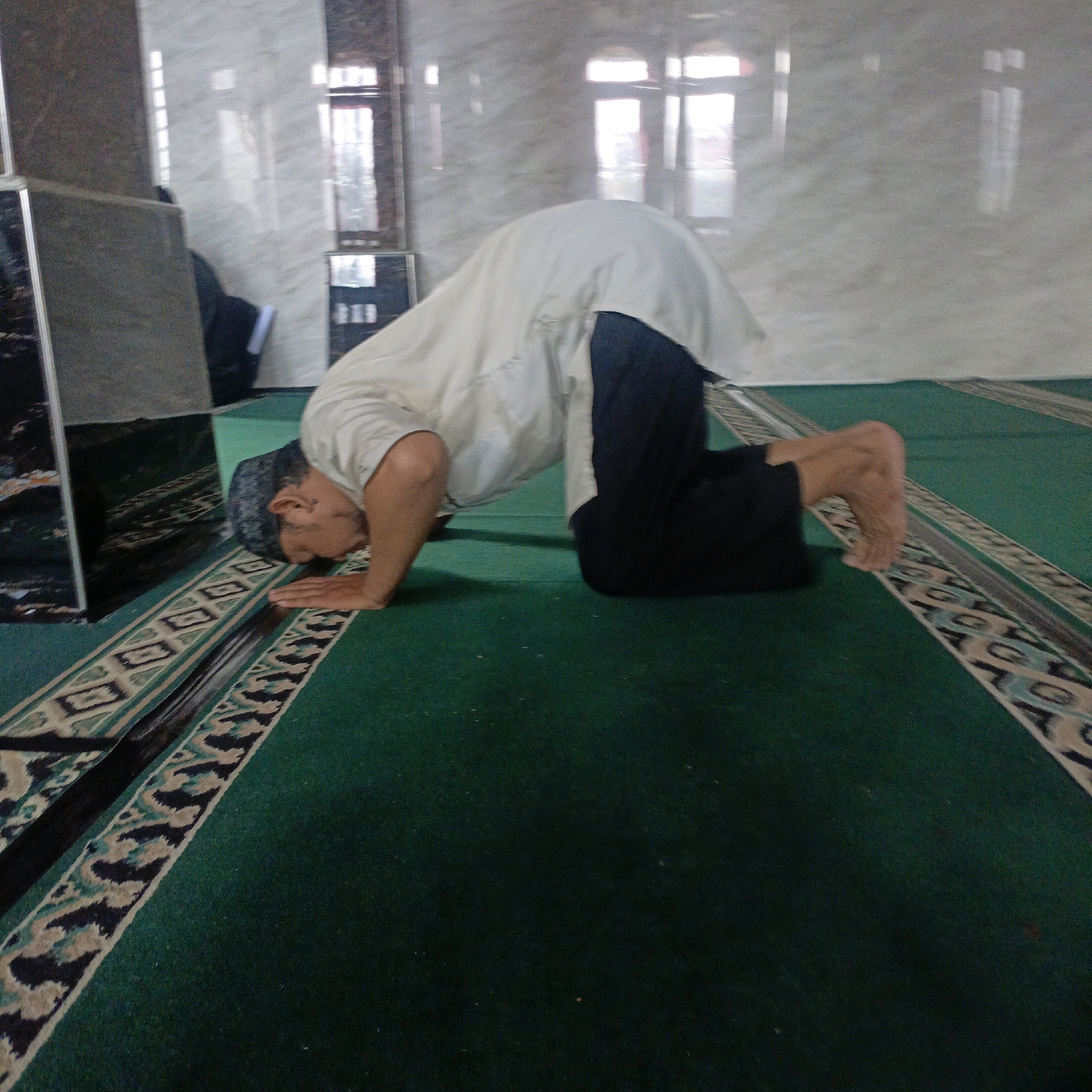 A man is performing Salat (Islamic prayer) in a mosque. He is in the Sujud position, with his forehead touching the ground. He is wearing a white shirt and dark pants. The mosque has green carpet with decorative patterns. The walls are made of marble. There are windows with wooden frames visible in the background. The image appears to be taken in Salatiga, Indonesia, based on the provided information.