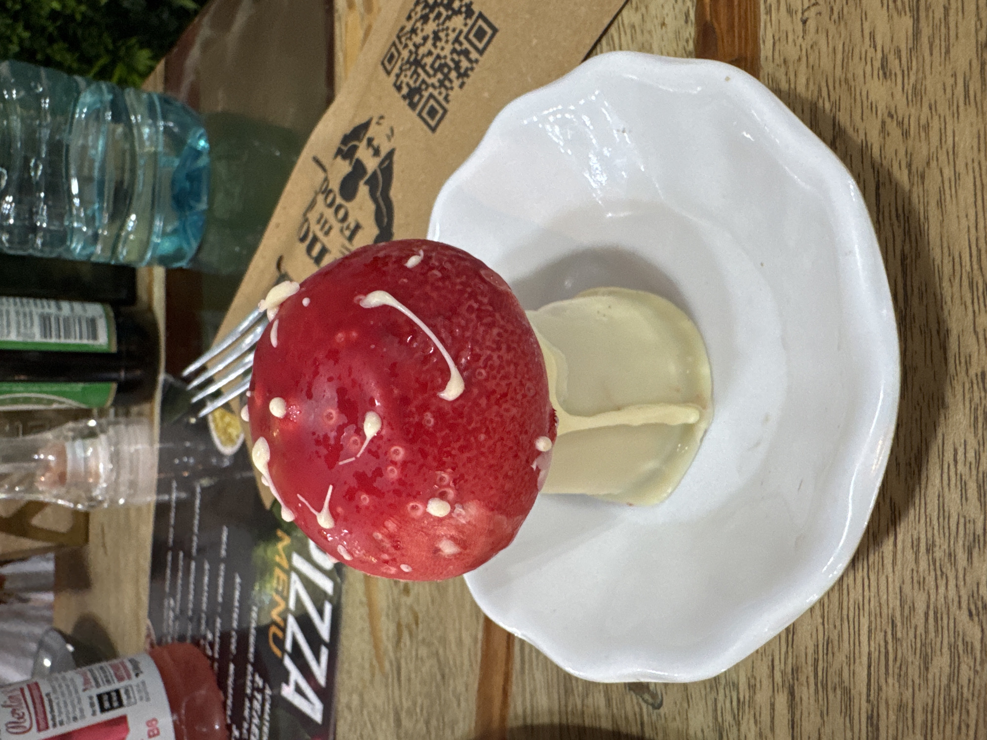 A top-down, close-up shot depicts a dessert resembling a mushroom on a white plate, placed on a wooden table. The "mushroom cap" is red with white markings, while the "stem" is a light, creamy color. A water bottle, part of a menu with the word "PIZZA" prominently displayed, a fork, and a bottle of seasoning are partially visible in the background. The scene is illuminated with an indoor light source. Based on the menu visible, the likely location is a restaurant serving pizza, somewhere in the city of Sofia, Bulgaria.
