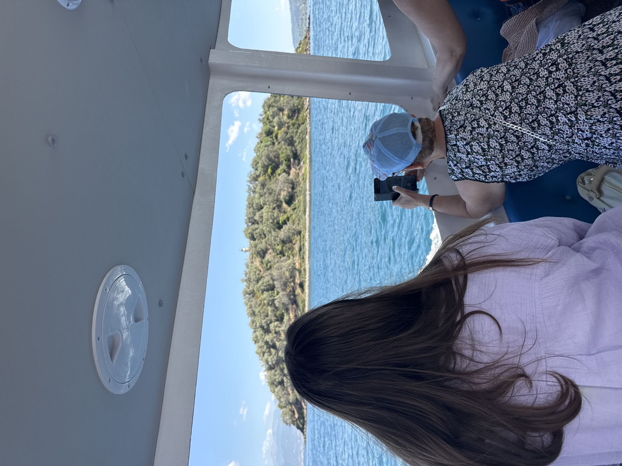A rear view shows two people inside a boat, looking out at a small, tree-covered island with a lighthouse in the distance, located in the waters off the coast of Greece. The person in the foreground, on the left, has long dark brown hair and wears a light purple shirt. To their right, a person wearing a blue baseball cap and a floral dress holds up a phone to take a picture of the island. The boat's interior is visible, with a white ceiling panel featuring a circular vent and a window framing the scenic view of the turquoise sea, the island, and a clear blue sky with scattered clouds.