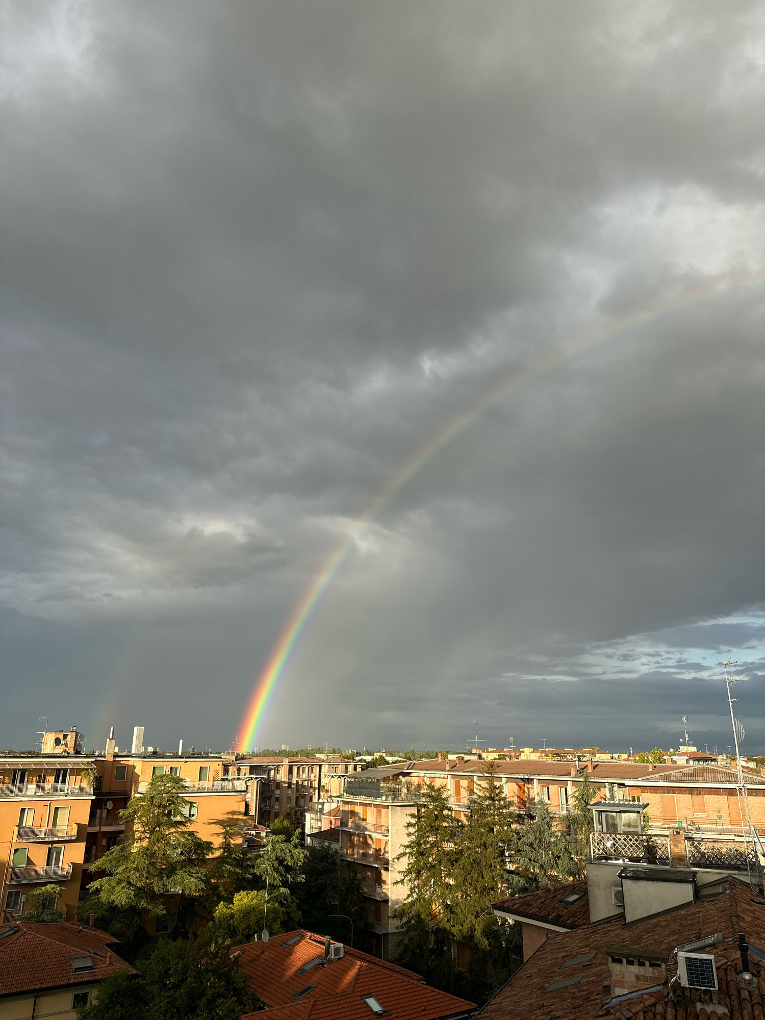 The image depicts a cityscape with a double rainbow arcing across a stormy, overcast sky. The scene is likely taken from an elevated vantage point, looking out over a densely populated urban area. Buildings, predominantly made of brick and painted in muted tones of orange and brown, are visible in the foreground and midground. Some buildings have balconies and trees are interspersed among them, some appearing to be evergreen. The roofs are predominantly tiled in red. In the distance, taller structures and a few antennas can be seen. The sky is dominated by dark, heavy clouds, with patches of lighter gray and some areas of blue peeking through. The light suggests either early morning or late afternoon, with the sun illuminating parts of the scene and the rainbow. The presence of a rainbow implies recent or ongoing precipitation. The overall impression is one of a city experiencing a break in the weather after a storm. The specific location is the city of Parma, Italy, as indicated by the architectural style and context of the image. There are no people or discernible actions visible in the image.