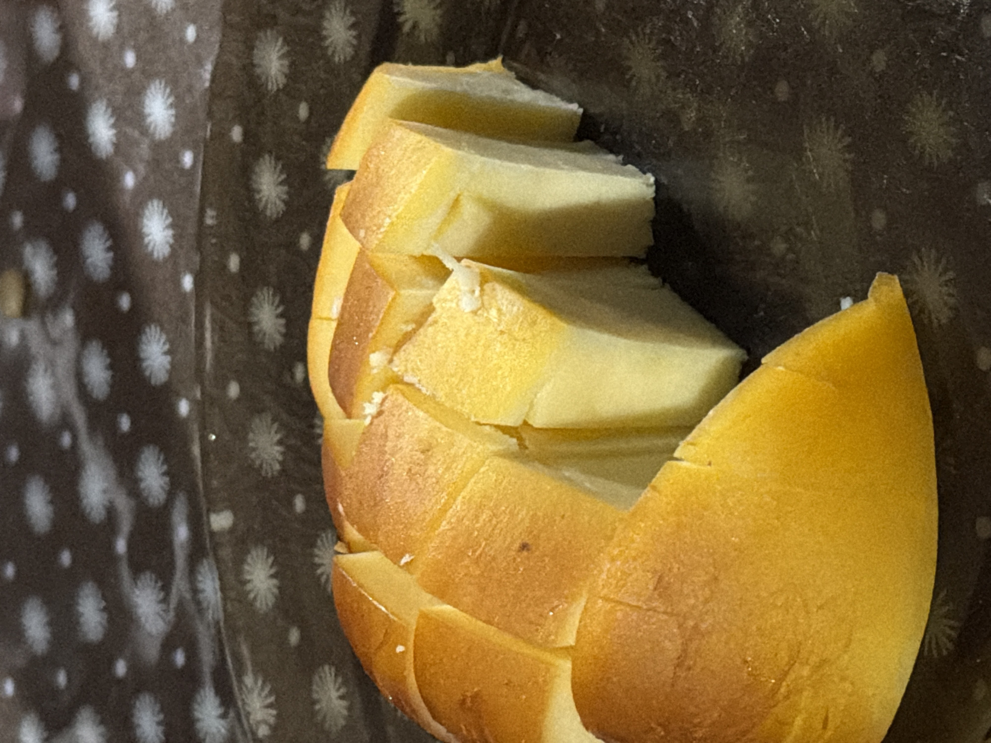 The image shows a close-up, overhead view of a golden-brown, sliced bread on a dark, patterned plate. The bread appears to be a sweet bread or cake, with a soft, light yellow interior. The top crust is lightly browned and slightly textured. The bread is cut into rectangular pieces, revealing the layered structure. The plate has a subtle, dark pattern of white star-like shapes and small dots. The background is out of focus, showing more of the same patterned plate and a hint of a darker surface. The lighting highlights the glossy sheen of the bread's crust. There are no people, actions, or interactions visible in this still life image. The location in the city of Sochi, Russia, is not discernible from the visual information provided.