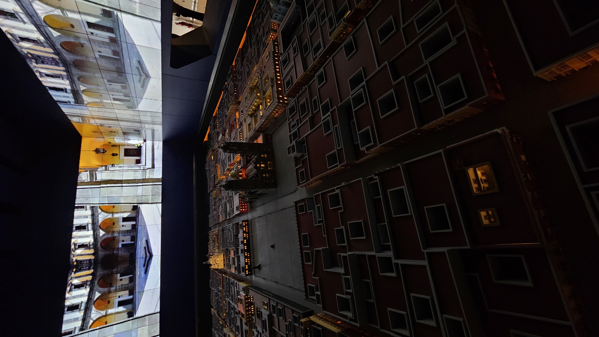The image depicts a museum display showcasing a miniaturized urban landscape, potentially representing the city of San Bernardino Tlaxcalancingo, Mexico. On the left side, a vertical arrangement of screens displays architectural scenes that may depict real locations within San Bernardino Tlaxcalancingo. These scenes feature archways, courtyards, and building facades in shades of white, gray, and yellow.
To the right, a detailed model city extends into the frame. The buildings are primarily constructed in a reddish-brown color with white trim around windows, presenting a grid-like street layout. Small lights illuminate some windows, suggesting activity within the miniature buildings. The scene is dimly lit overall, creating a dramatic effect.
No people are visible in the image. The scene focuses solely on the built environment, emphasizing the architecture and urban planning represented in the miniature model and corresponding video screens.