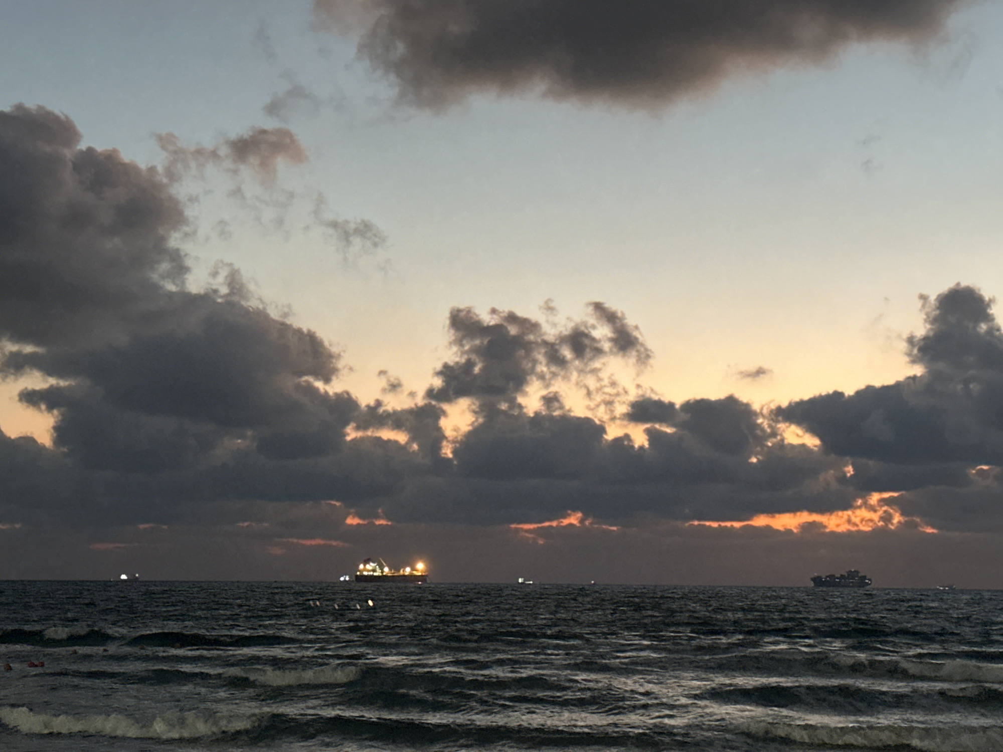 The image depicts a seascape at dusk, with a horizon line dividing the dark, choppy ocean from a sky filled with dramatic clouds. The sky transitions from a pale blue at the top to hues of orange and yellow near the horizon, indicating the setting sun. Large, dark clouds dominate the upper and middle sections of the sky, with some lighter patches where the sunset light breaks through.
On the water, several ships are visible in the distance. One prominent ship, likely a cargo vessel, is illuminated by lights and is positioned to the right of the frame. Other smaller lights suggest additional vessels further out. The waves are moderate, with whitecaps visible, suggesting a breezy or slightly rough sea. The foreground shows the cresting of waves as they approach the shore.
The scene is set at Kiryat Yam, Israel, at dusk. The overall mood is somewhat somber due to the dark clouds and the dimming light, yet the warm glow of the sunset provides a touch of beauty and tranquility. There are no visible text elements in the image.